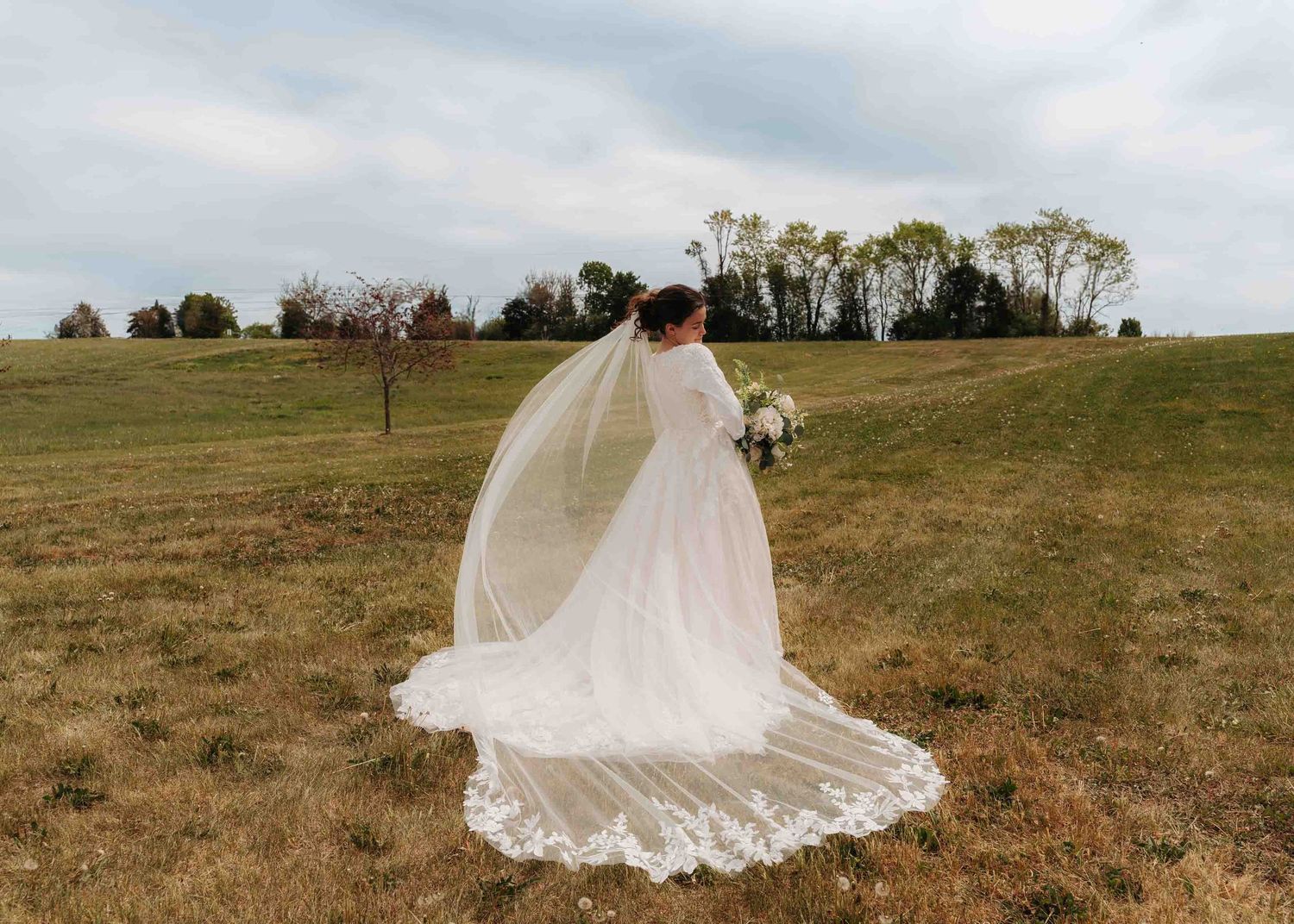 Bride in a flowing white gown with lace-trimmed veil walking across a scenic green countryside.
