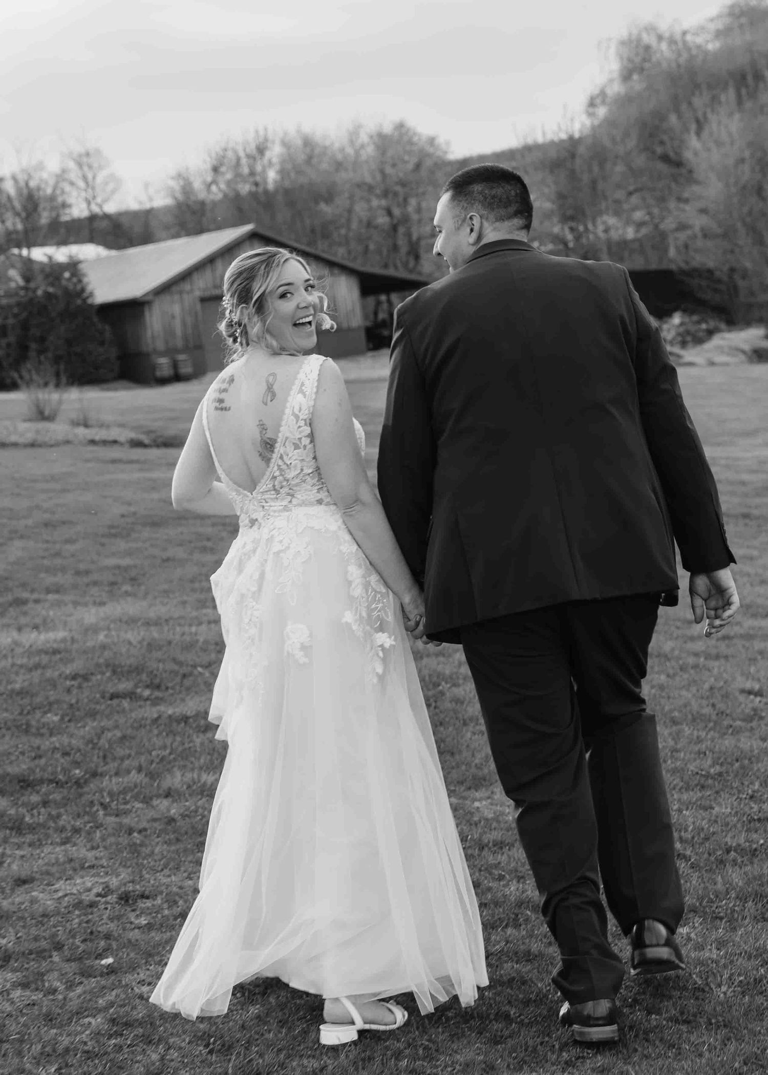 Black and white photo of laughing bride and groom walking at rustic farm wedding.