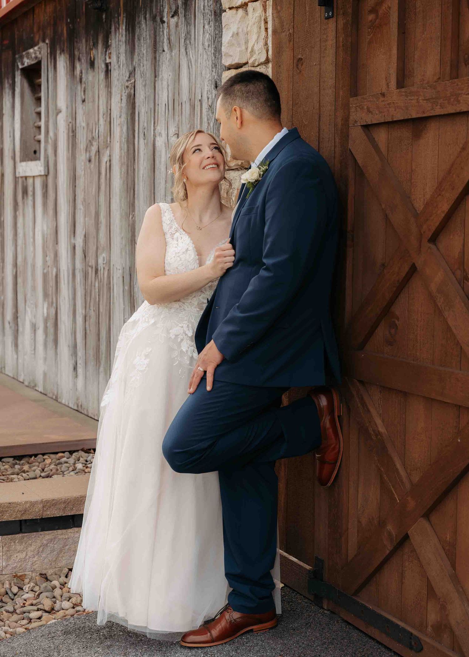 Smiling bride and groom share an intimate moment leaning against rustic barn wooden doors.