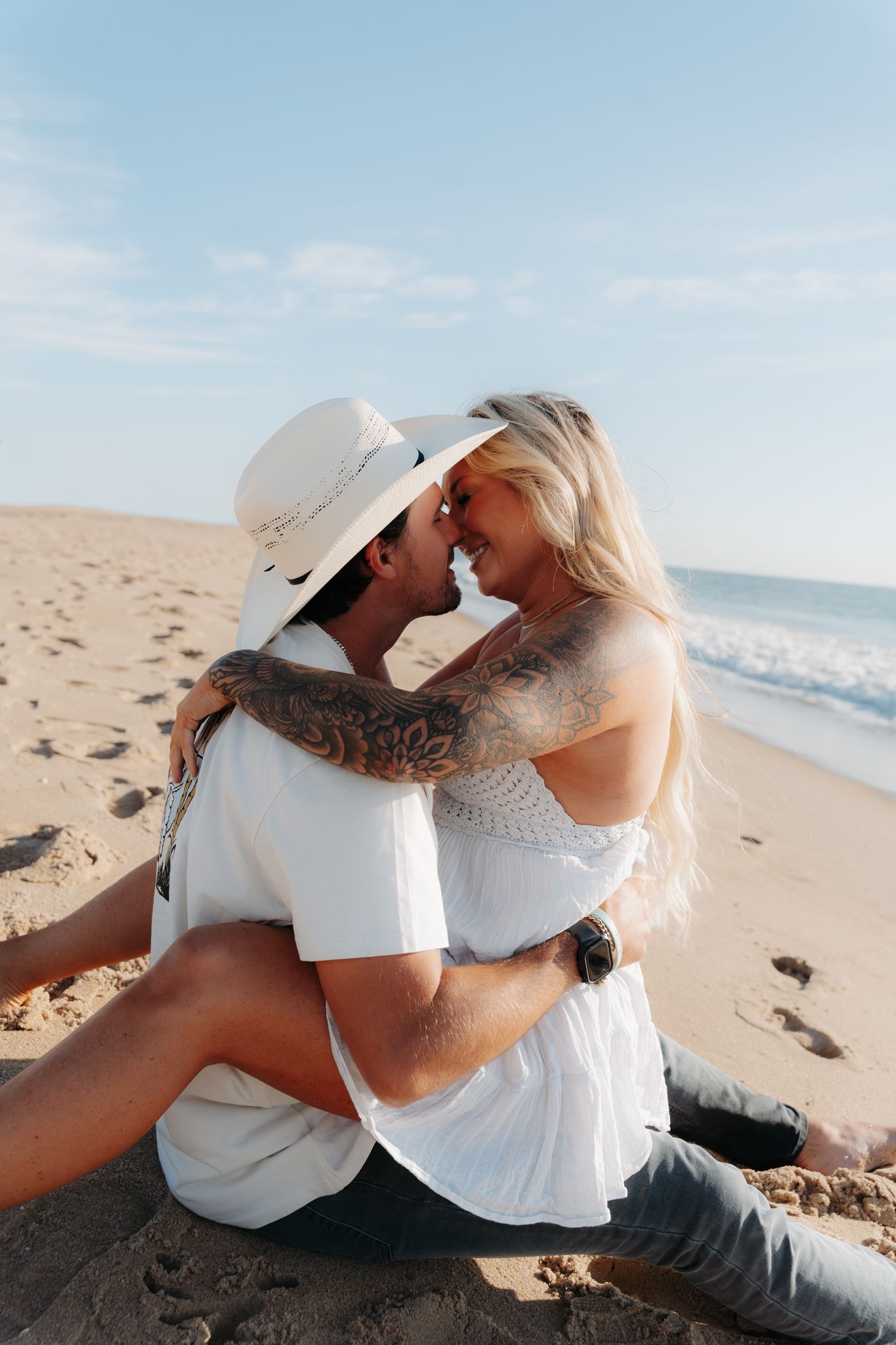 Romantic couple embracing on sandy beach, tattooed man in white hat and woman in white dress sharing a kiss.