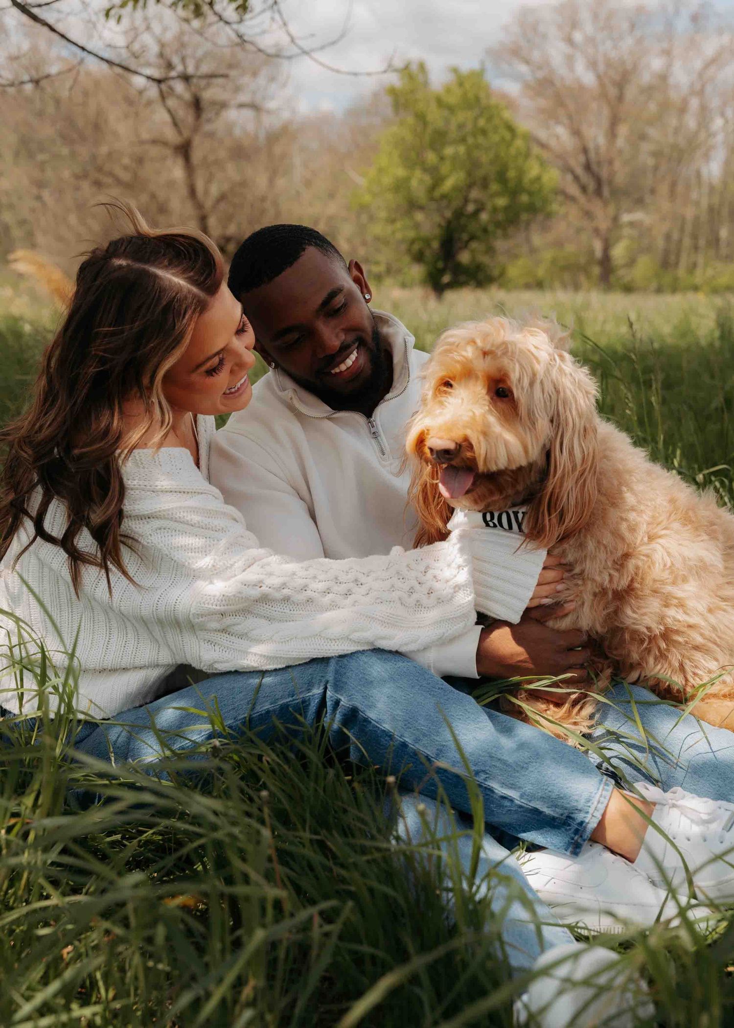 Smiling interracial couple sitting in grass with their fluffy golden doodle dog.