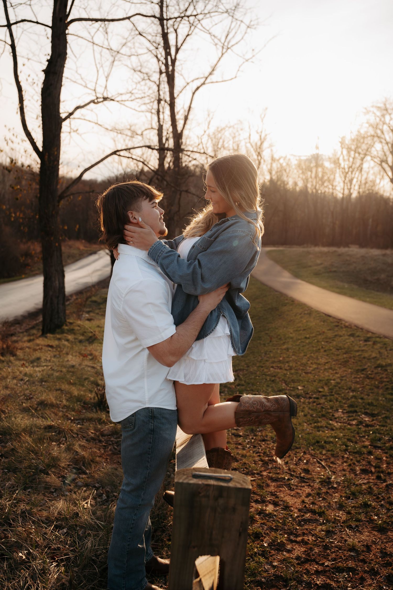 Couple embracing at golden hour, woman lifted onto a fence post along a rural road.