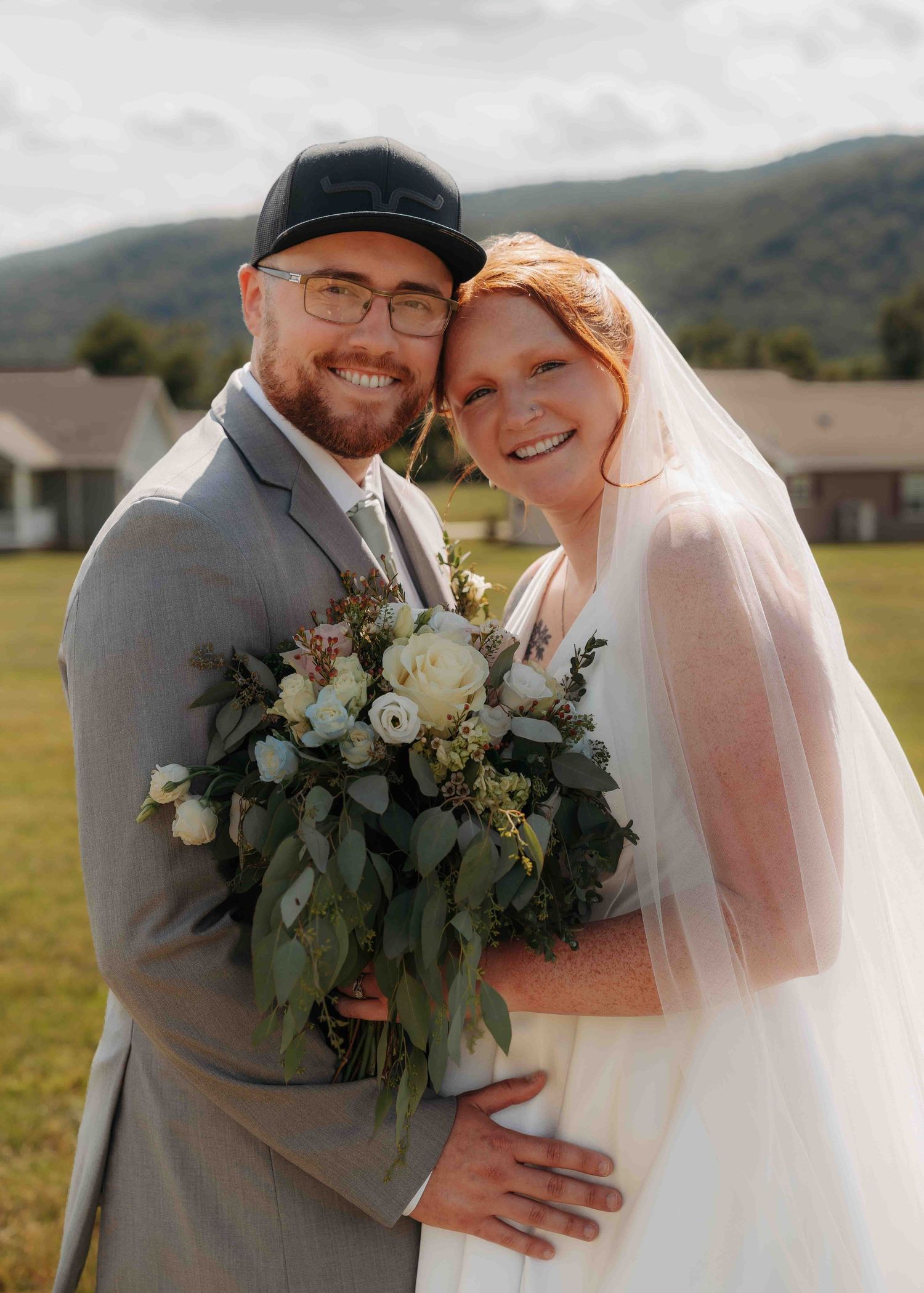 Smiling bride and groom pose together outdoors holding white and greenery bouquet with mountain scenery behind them.