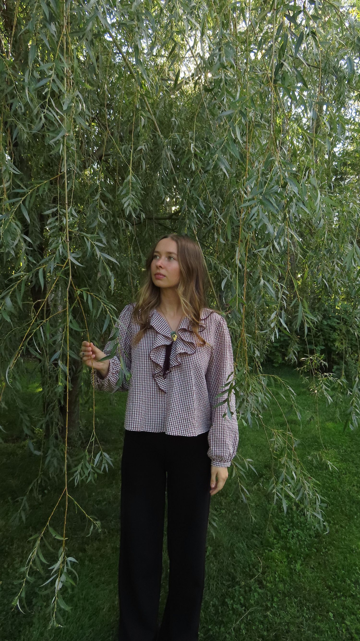 Young woman in gingham ruffle blouse and black wide-leg pants standing under weeping willow tree outdoors.