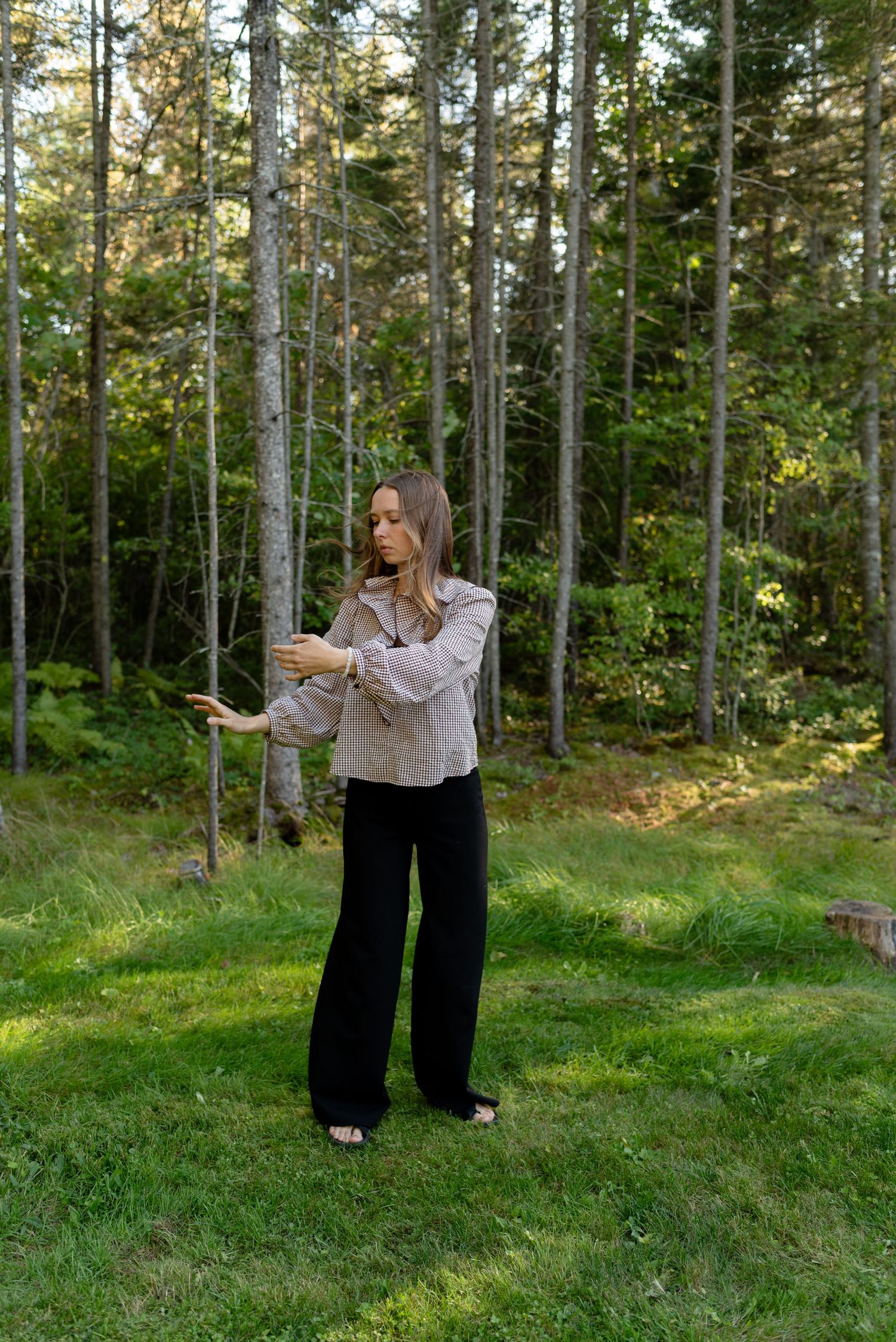 Woman in gingham blouse posing with arms extended in grassy clearing surrounded by pine forest.