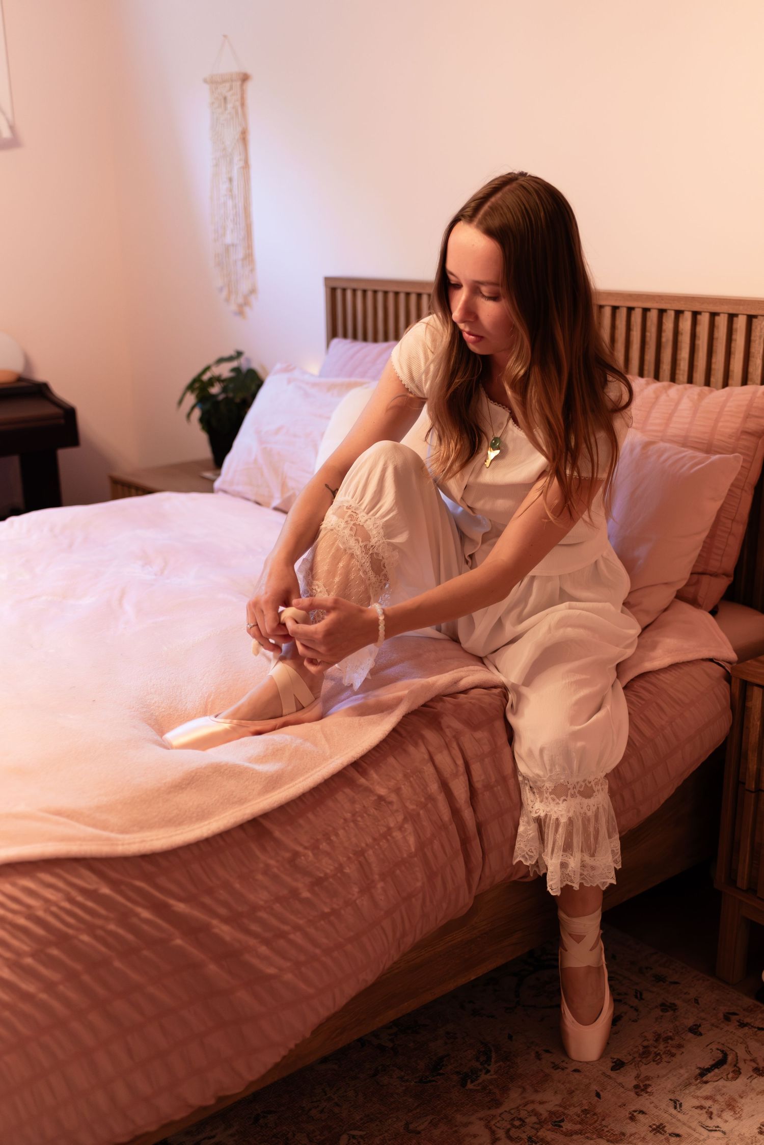 Young woman in white dress sitting on bed tying ballet pointe shoes in softly lit pink bedroom.
