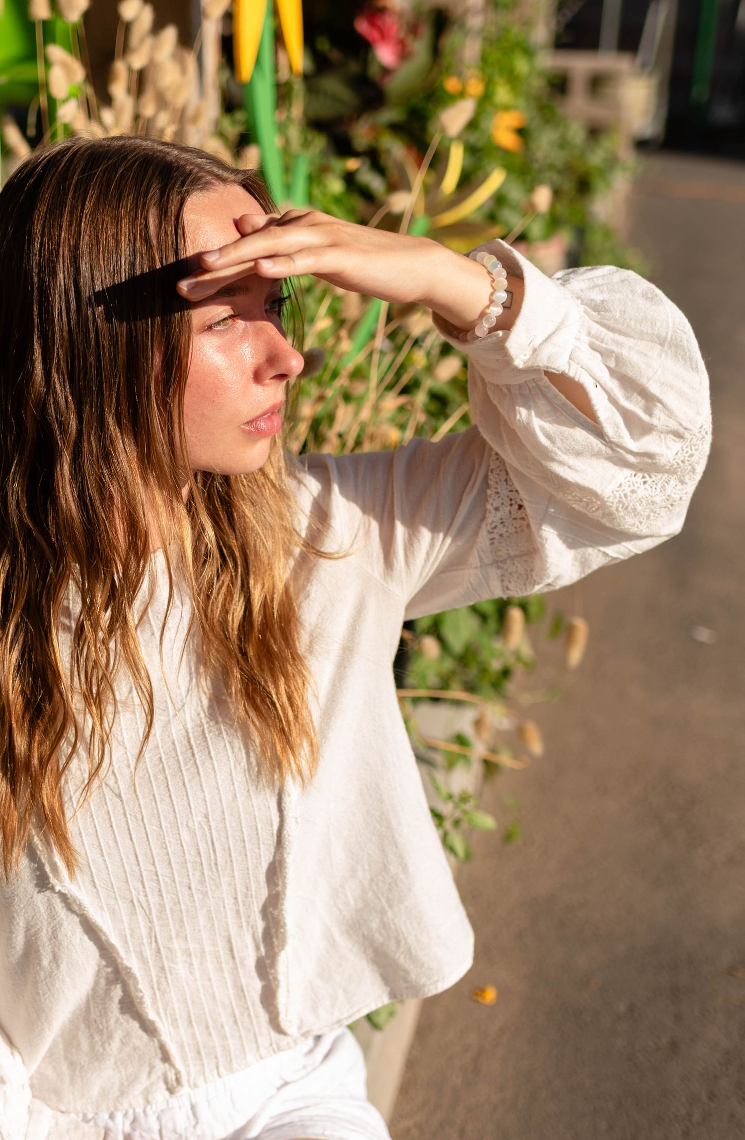 Woman in white lace blouse shielding her eyes from bright sunlight near lush tropical greenery.