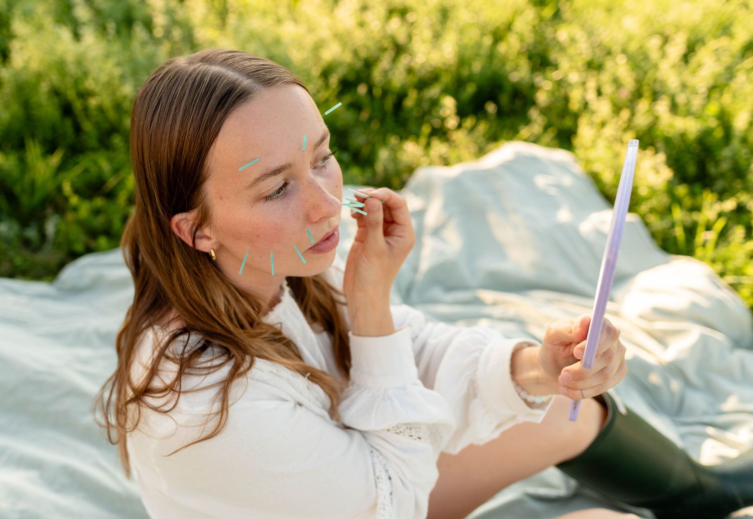 Young woman applying lip color using handheld mirror while sitting on blanket outdoors in sunlight.