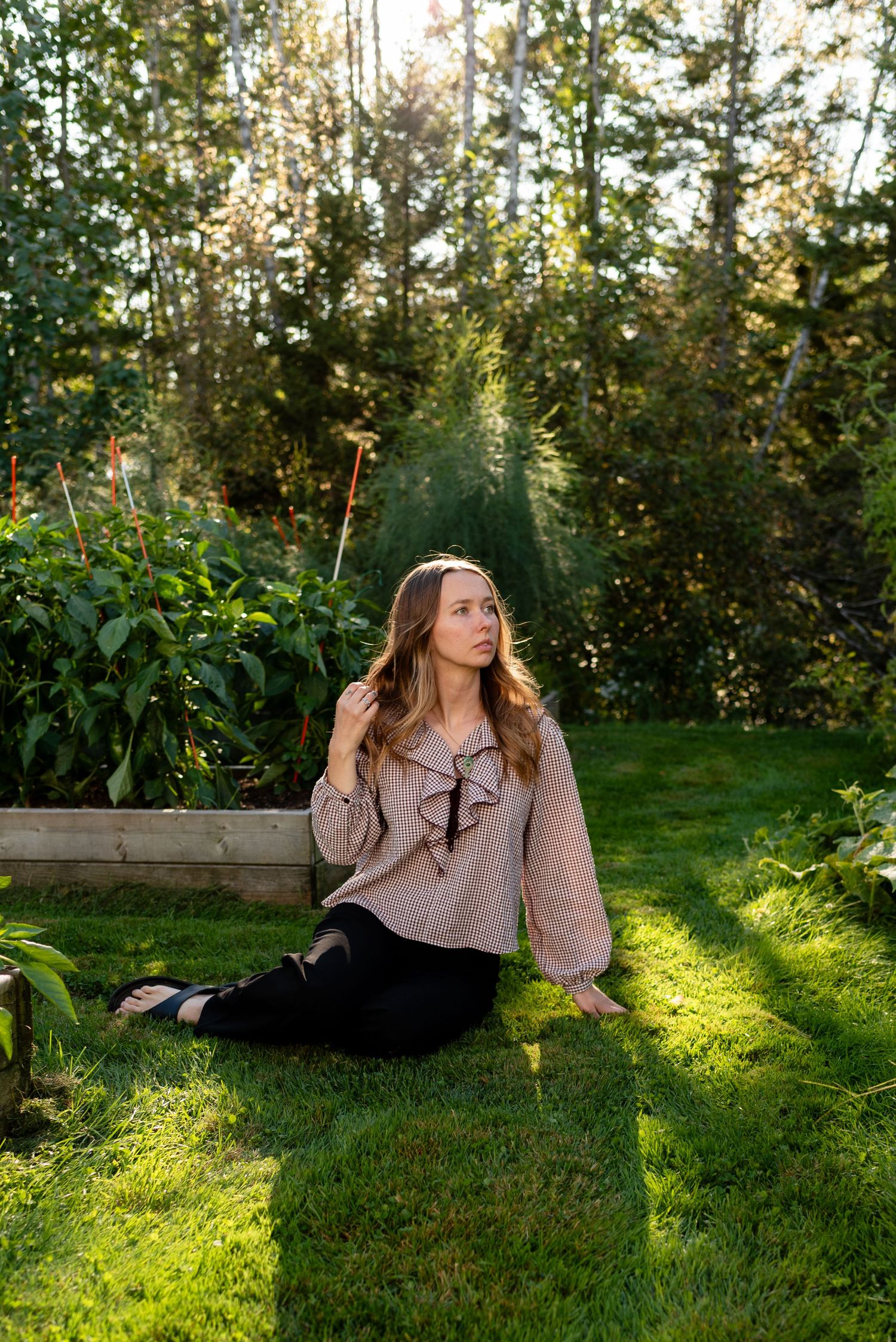 Woman in taupe ruffled blouse sitting on grass beside raised garden beds in dappled sunlight.