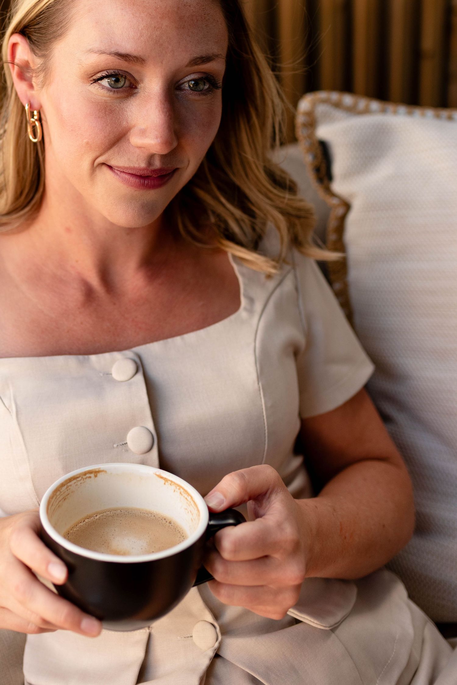Smiling blonde woman in beige dress holding a dark coffee cup while relaxing on a cushioned seat.