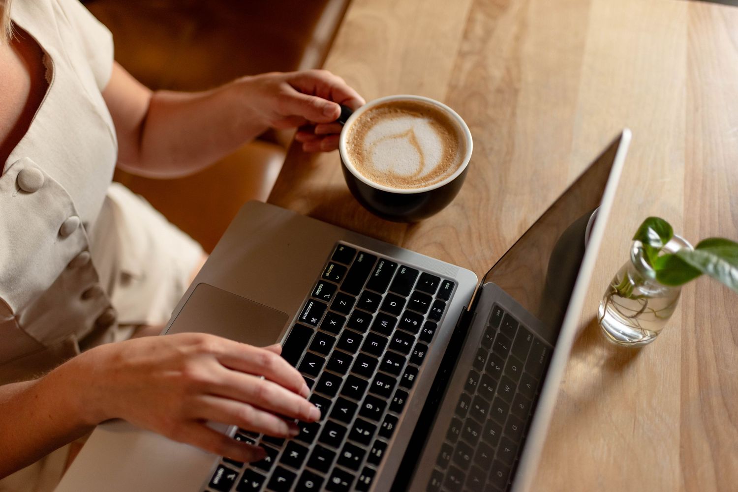 Woman typing on laptop while holding a latte art coffee at a wooden cafe table.