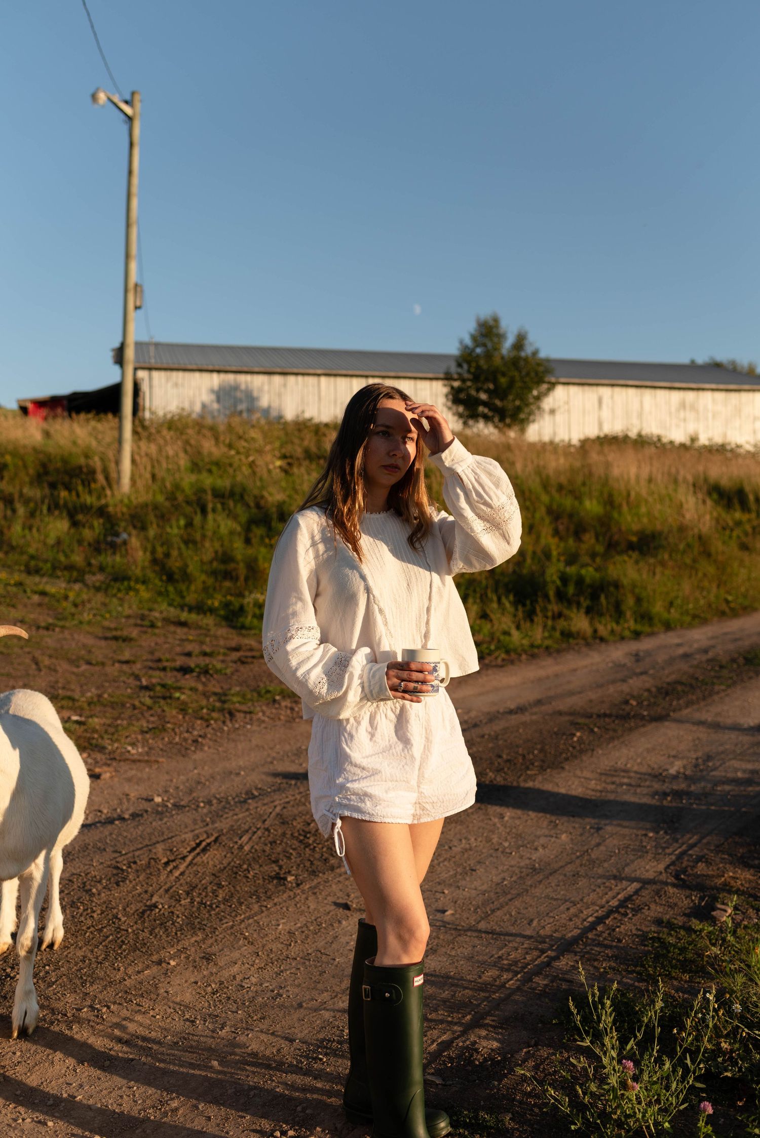 Woman in white shorts set and green boots shielding eyes from sun on farm road.
