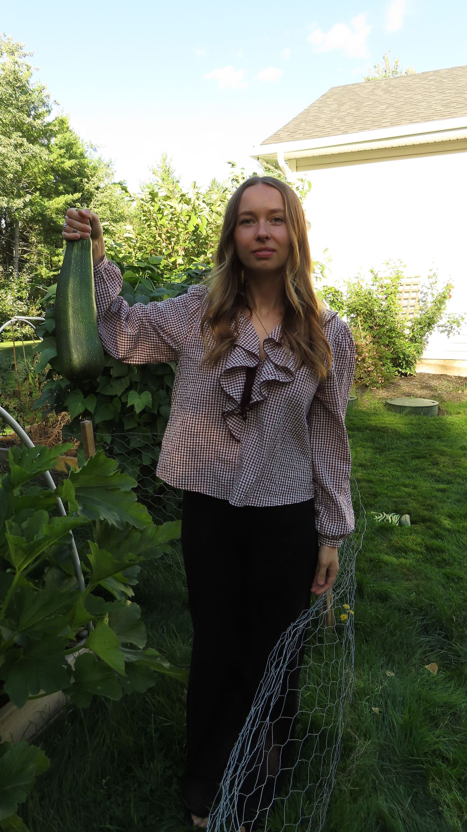 Woman in gingham ruffle top holding a large zucchini harvested from her backyard garden.