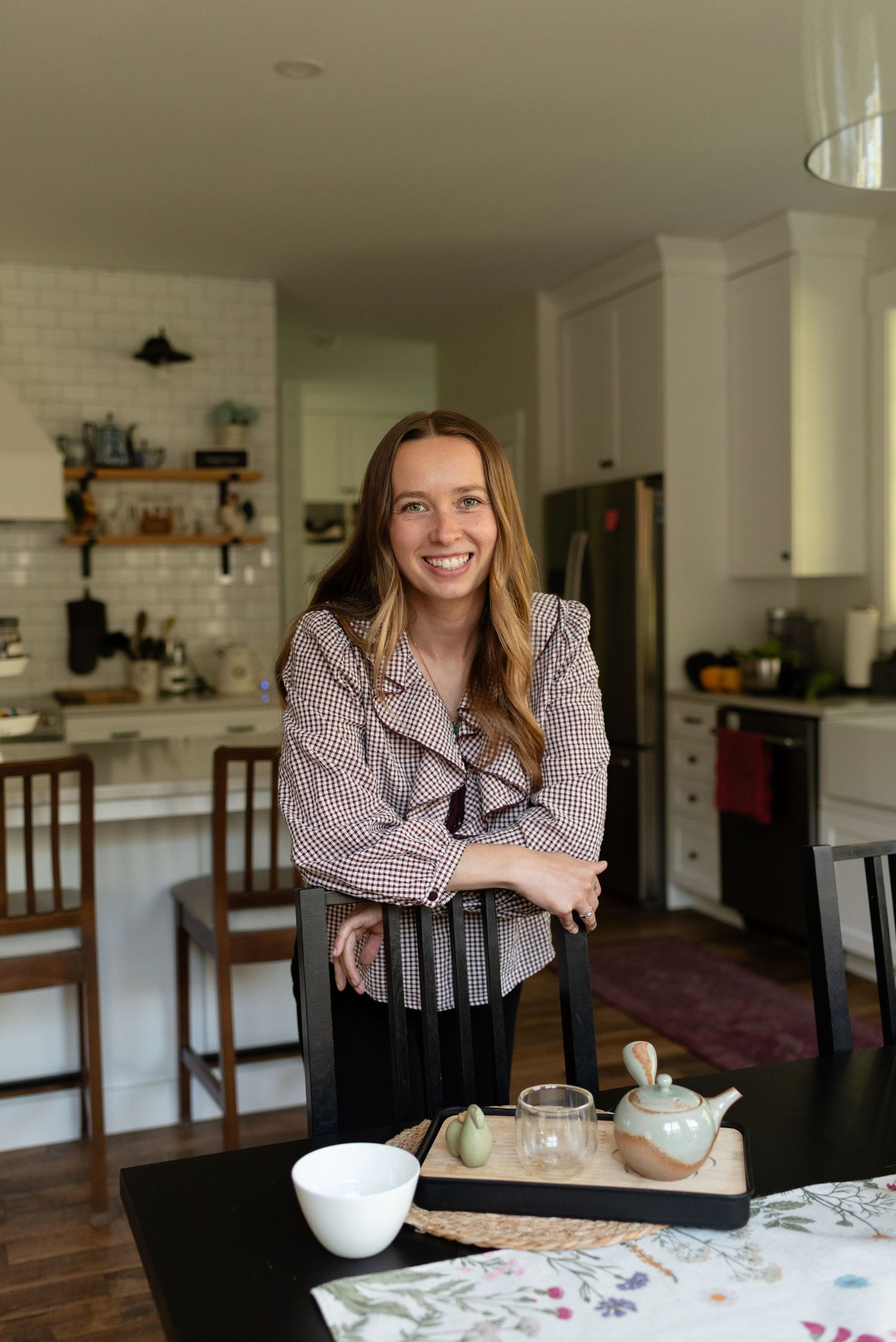 Smiling young woman in gingham blouse leaning on chair at dining table with tea set in bright kitchen.