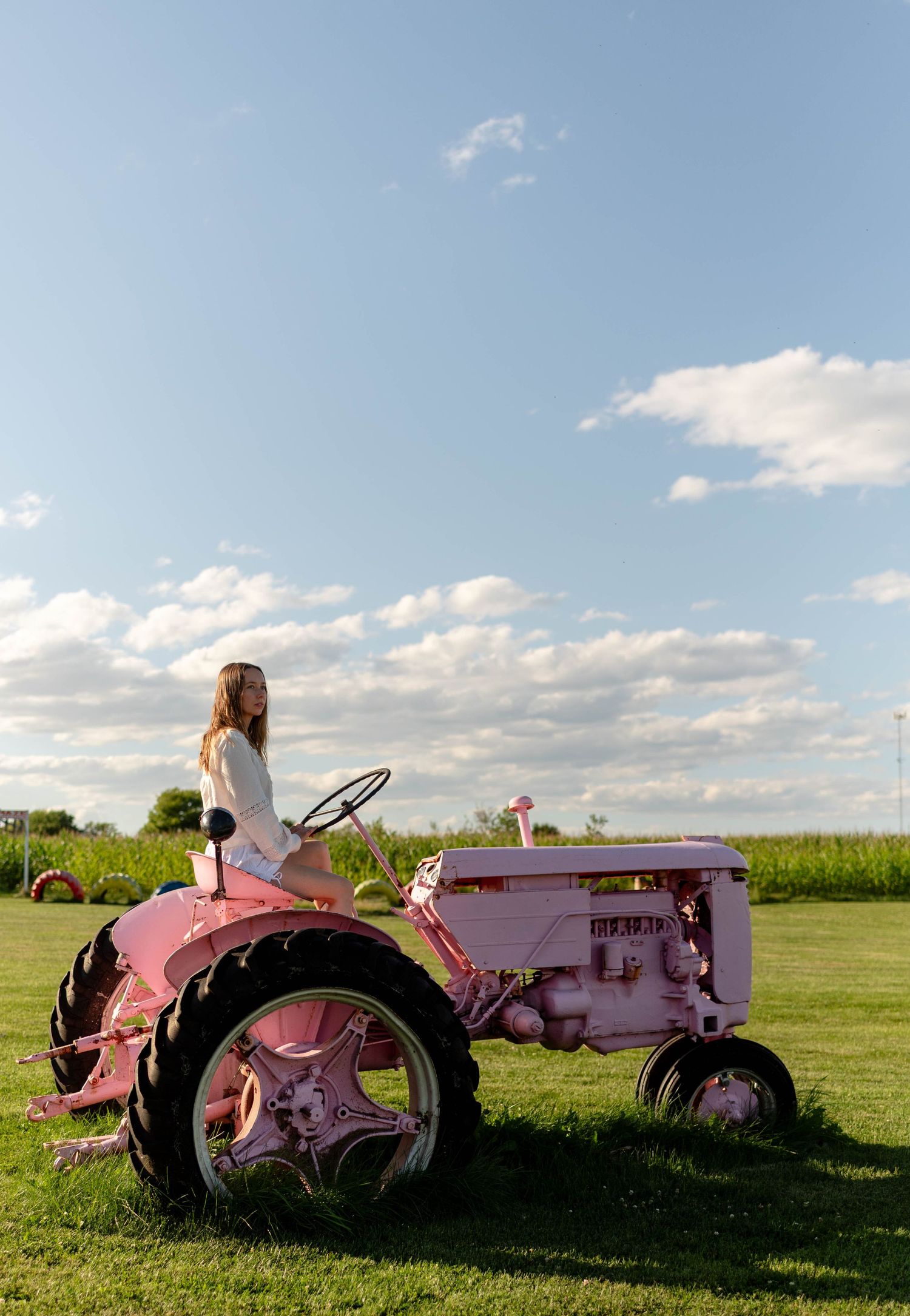 Woman in white outfit sitting atop a vintage pink tractor in an open green field under blue sky.