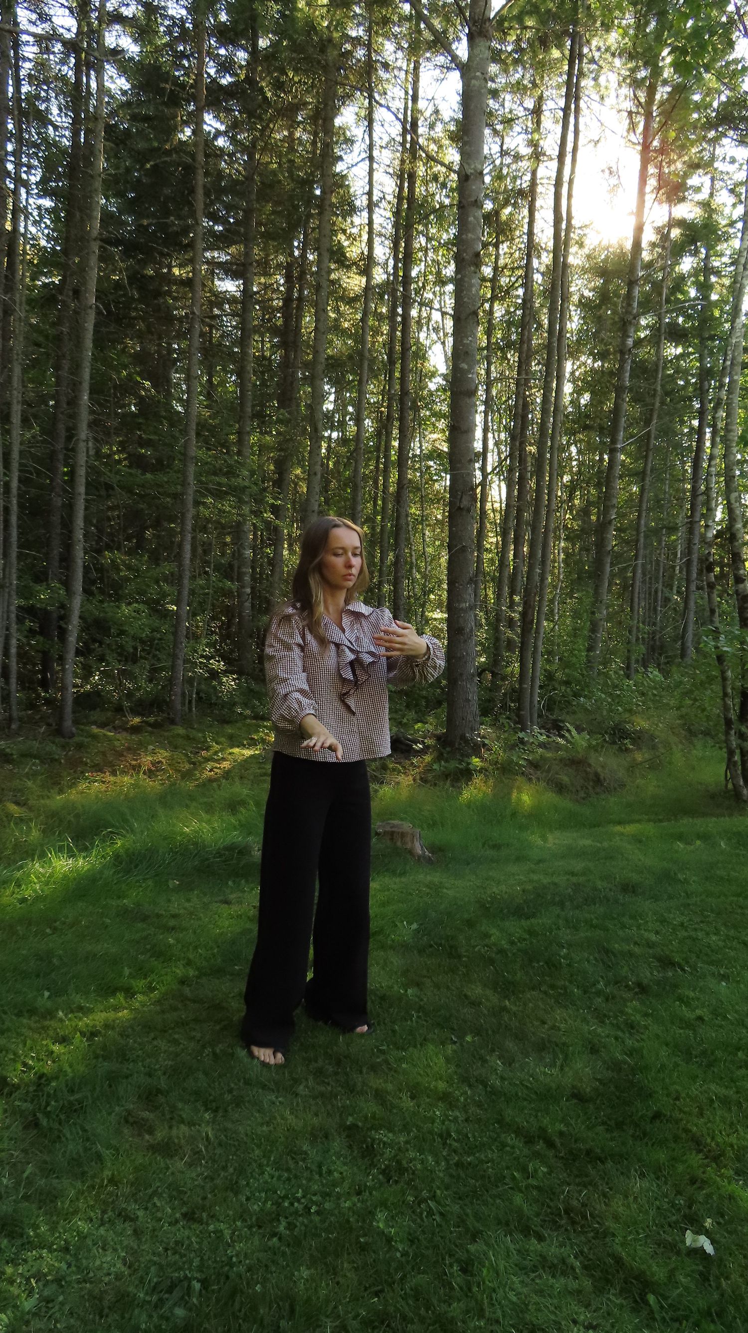 Woman in gingham blouse and black wide-leg pants standing in sunlit pine forest clearing.