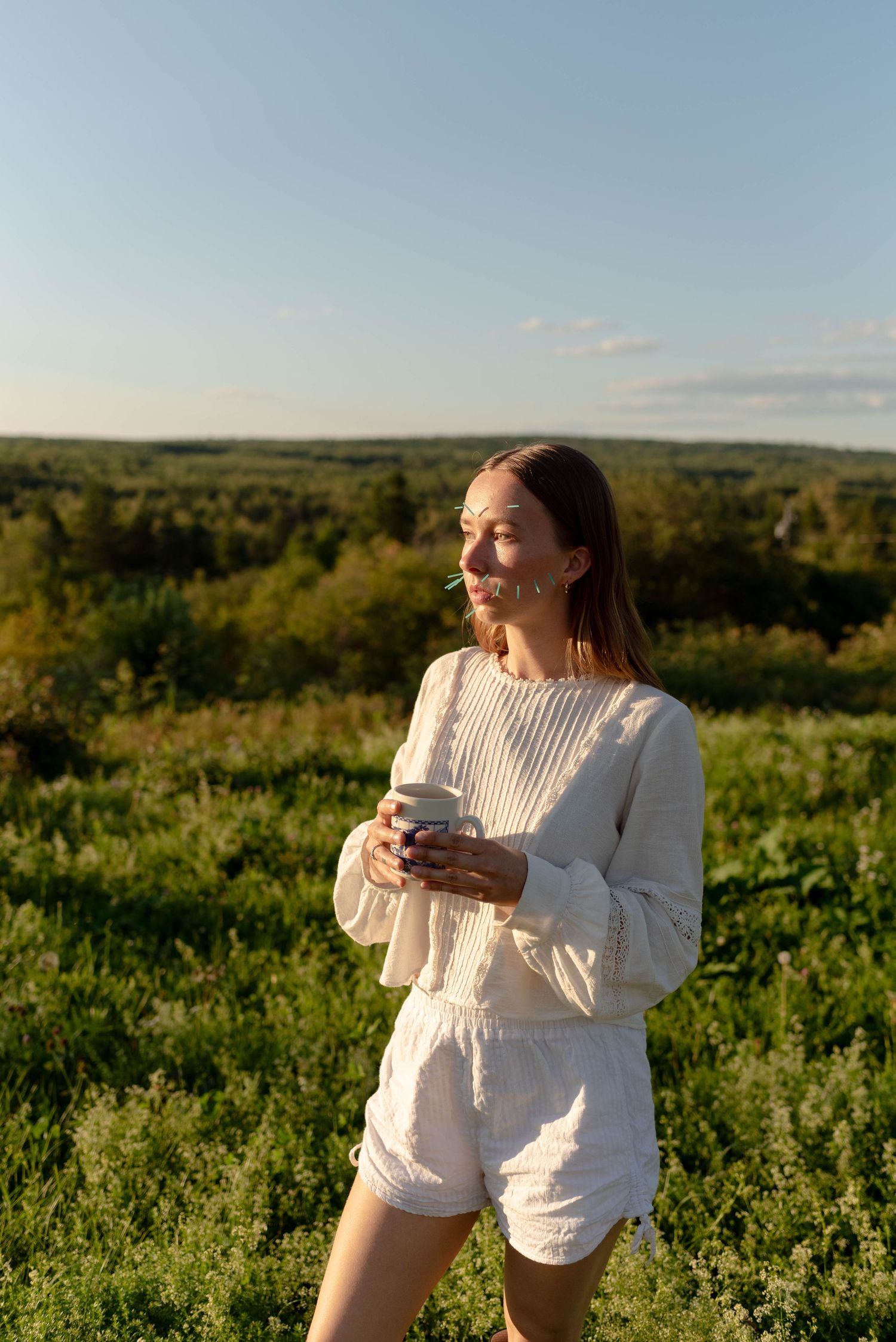 Woman in white matching set holding a small cup standing in a green field at golden hour sunset.