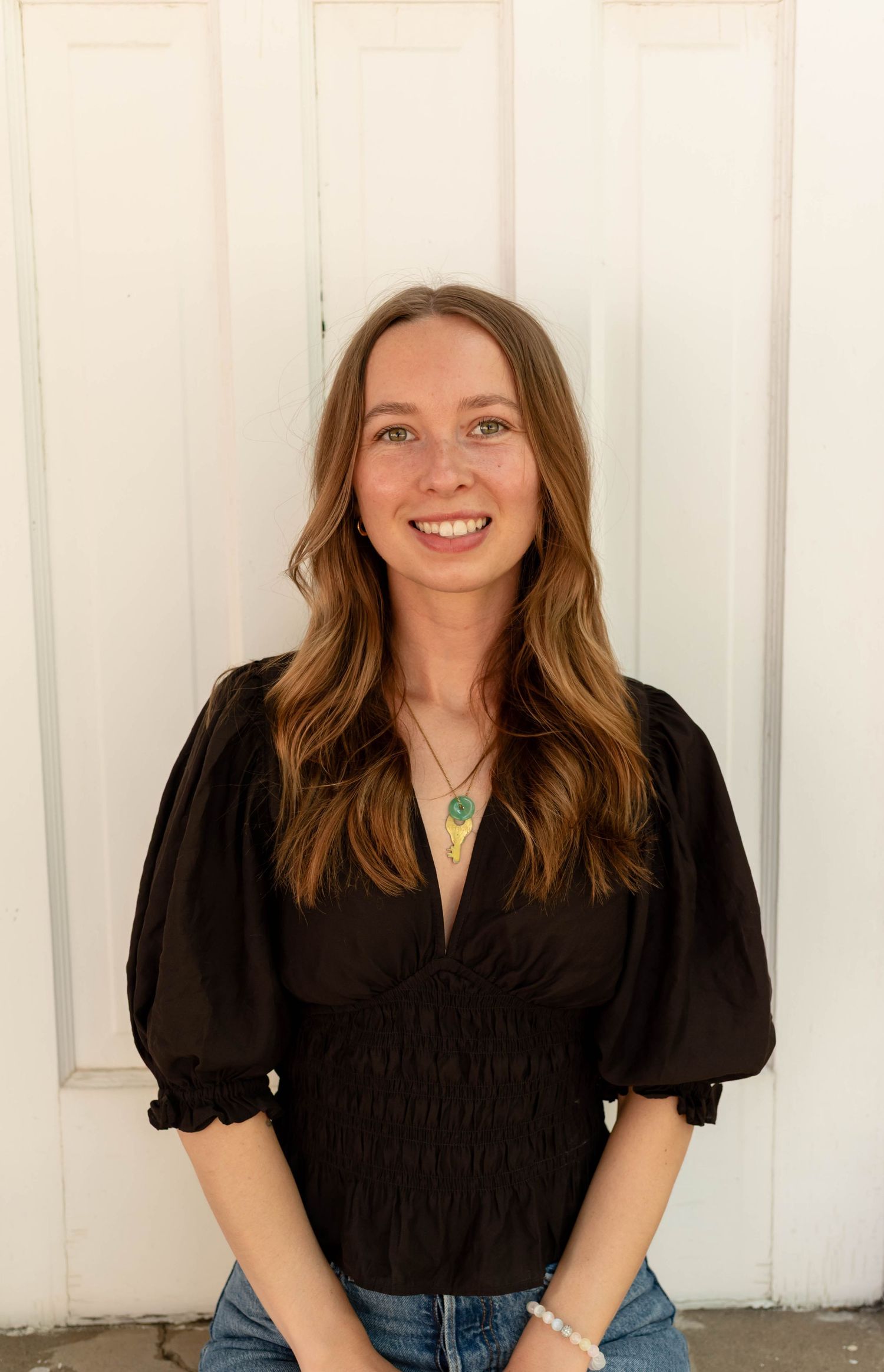 Smiling woman in black puff-sleeve top with green crystal pendant standing by white wall.