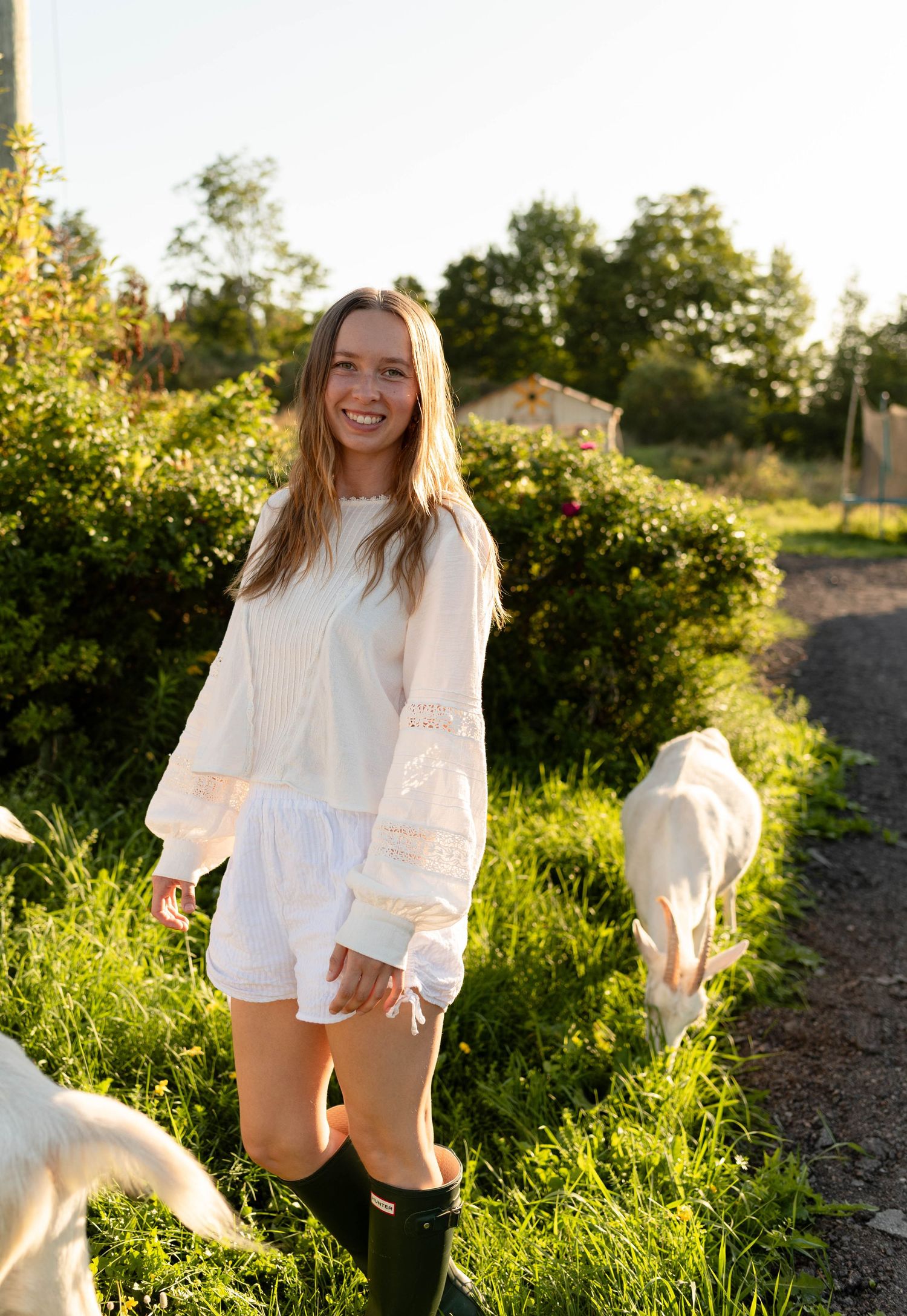 Smiling woman in white lace top and shorts standing in a sunlit garden path beside grazing goats.