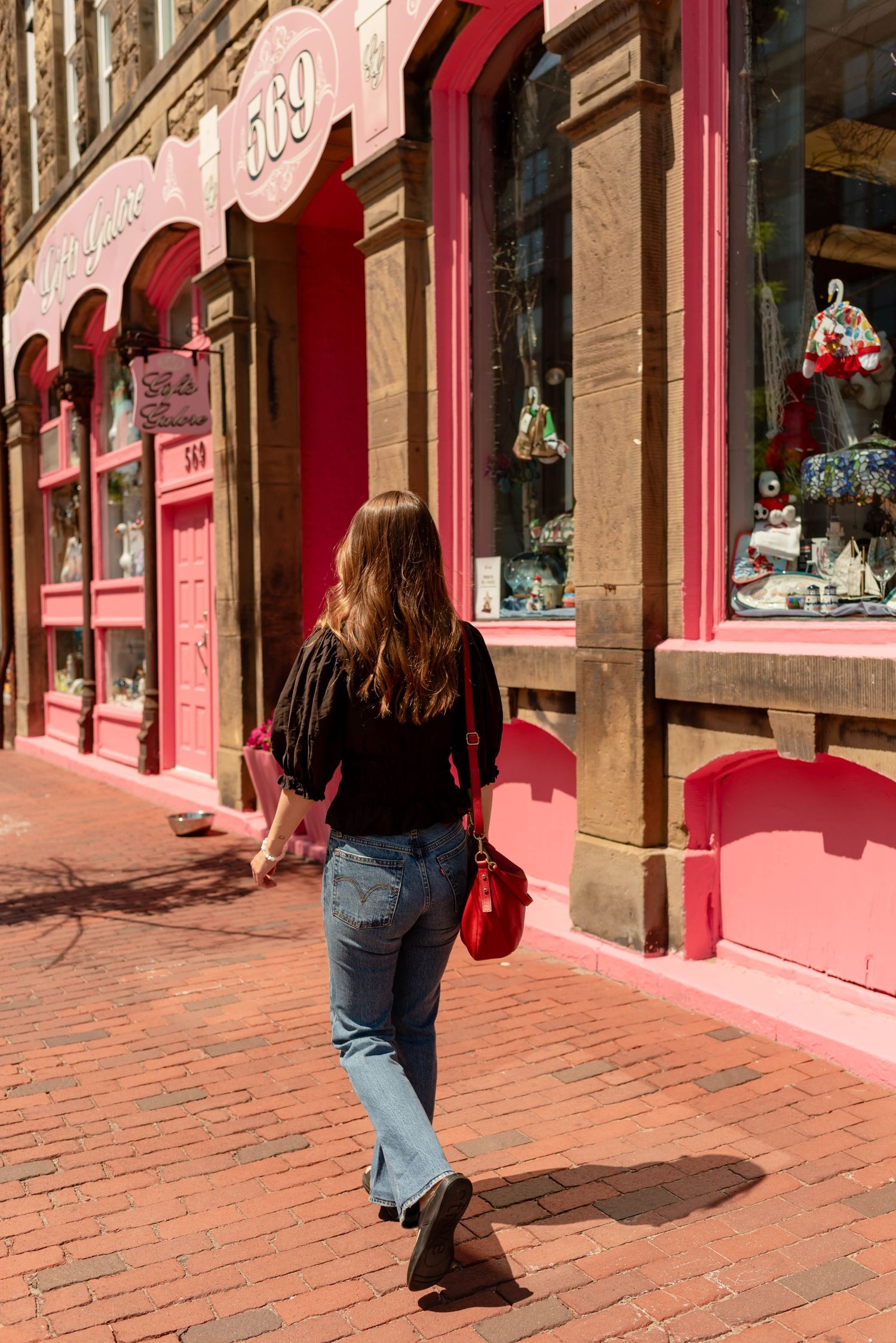 Woman in black top and jeans walking past pink storefront on brick sidewalk in historic district.
