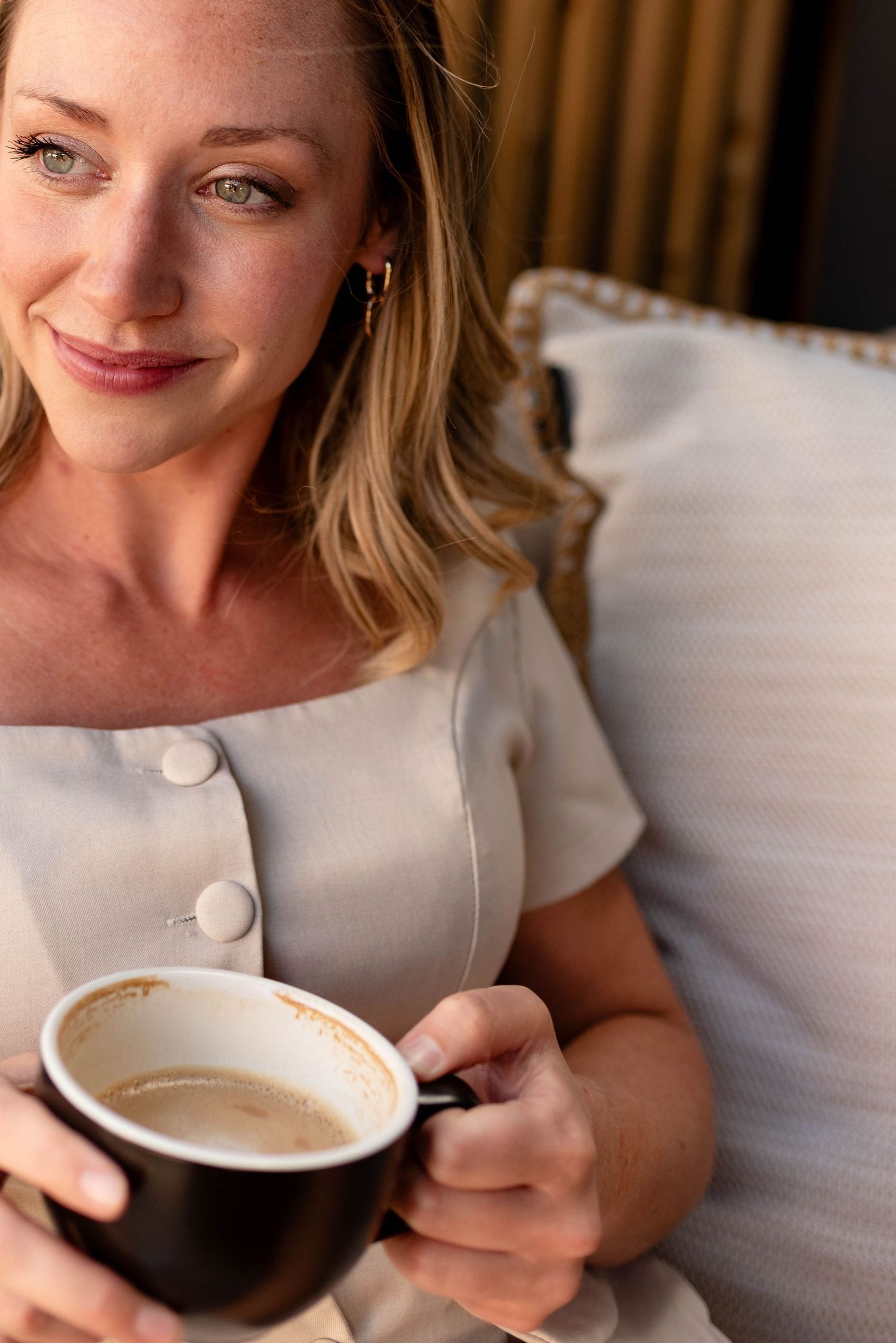 Elegant blonde woman in cream dress gazing softly while holding a black coffee cup indoors.