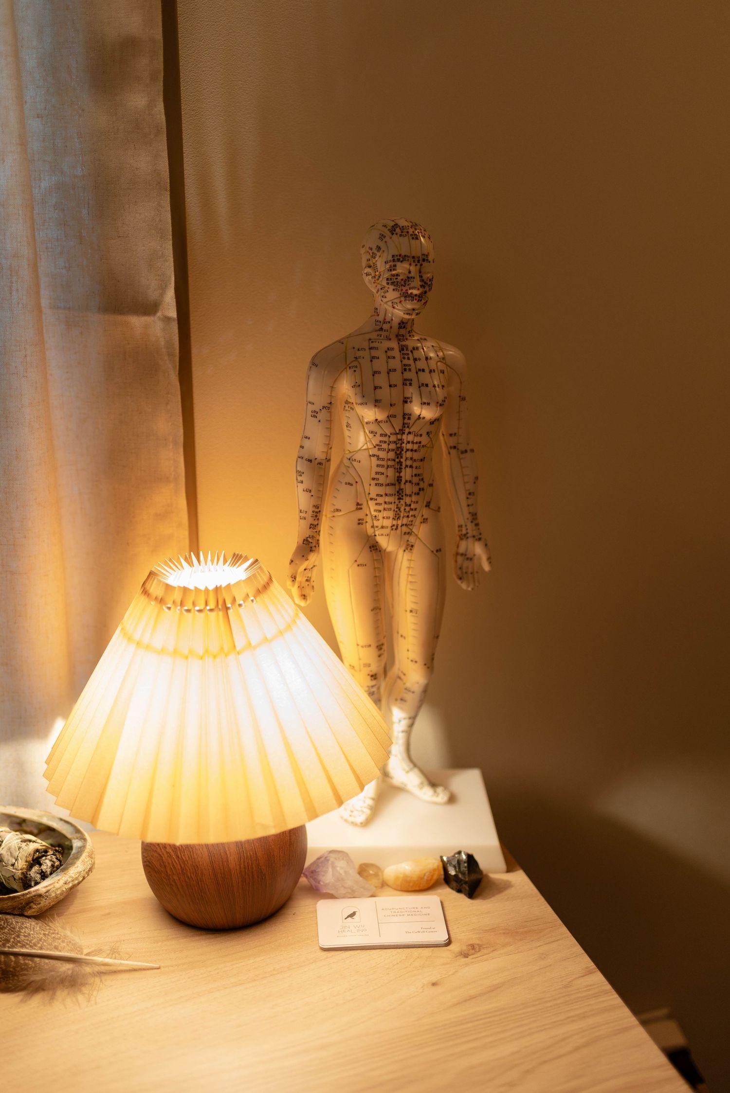 Acupuncture point model figurine displayed beside glowing lamp and crystals on wooden bedside table.