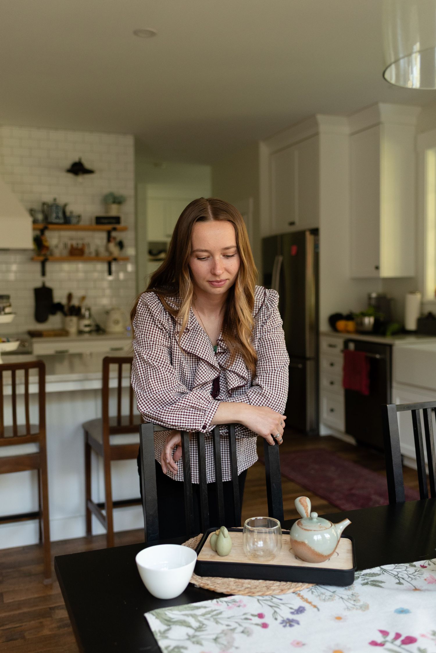 Young woman in plaid ruffled blouse standing at dining table with ceramic tea set indoors.