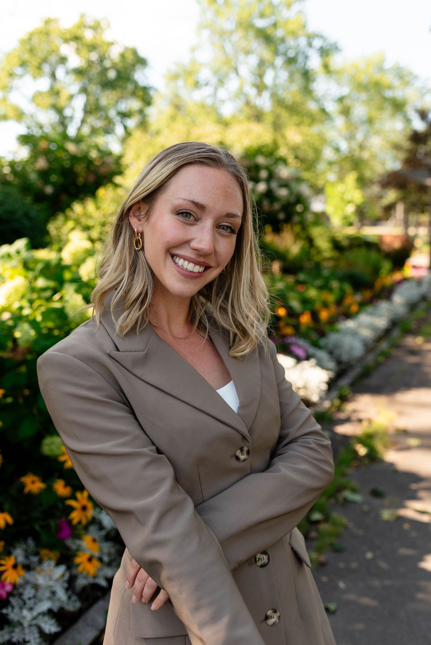 Smiling blonde woman in beige blazer with gold buttons standing in a sunny garden.