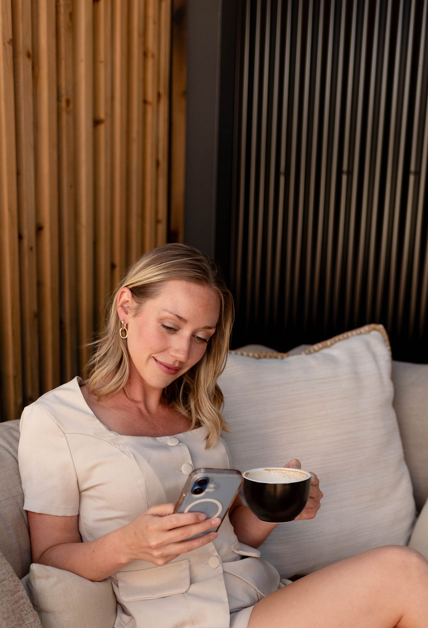 Blonde woman in beige dress using smartphone while holding coffee cup on sofa.
