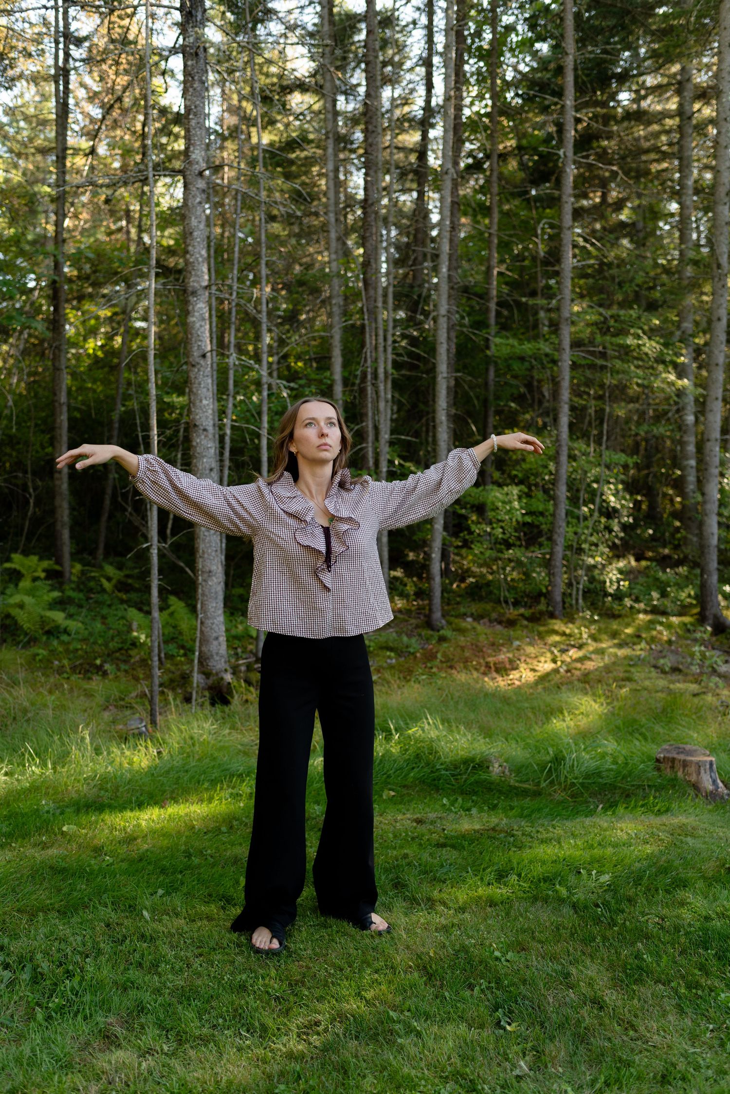 Woman in linen blouse with arms outstretched standing on grass in front of pine forest.