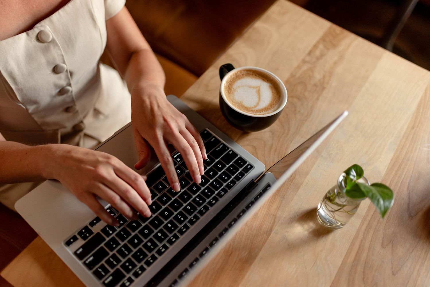 Overhead view of woman typing on laptop with latte coffee on wooden desk.
