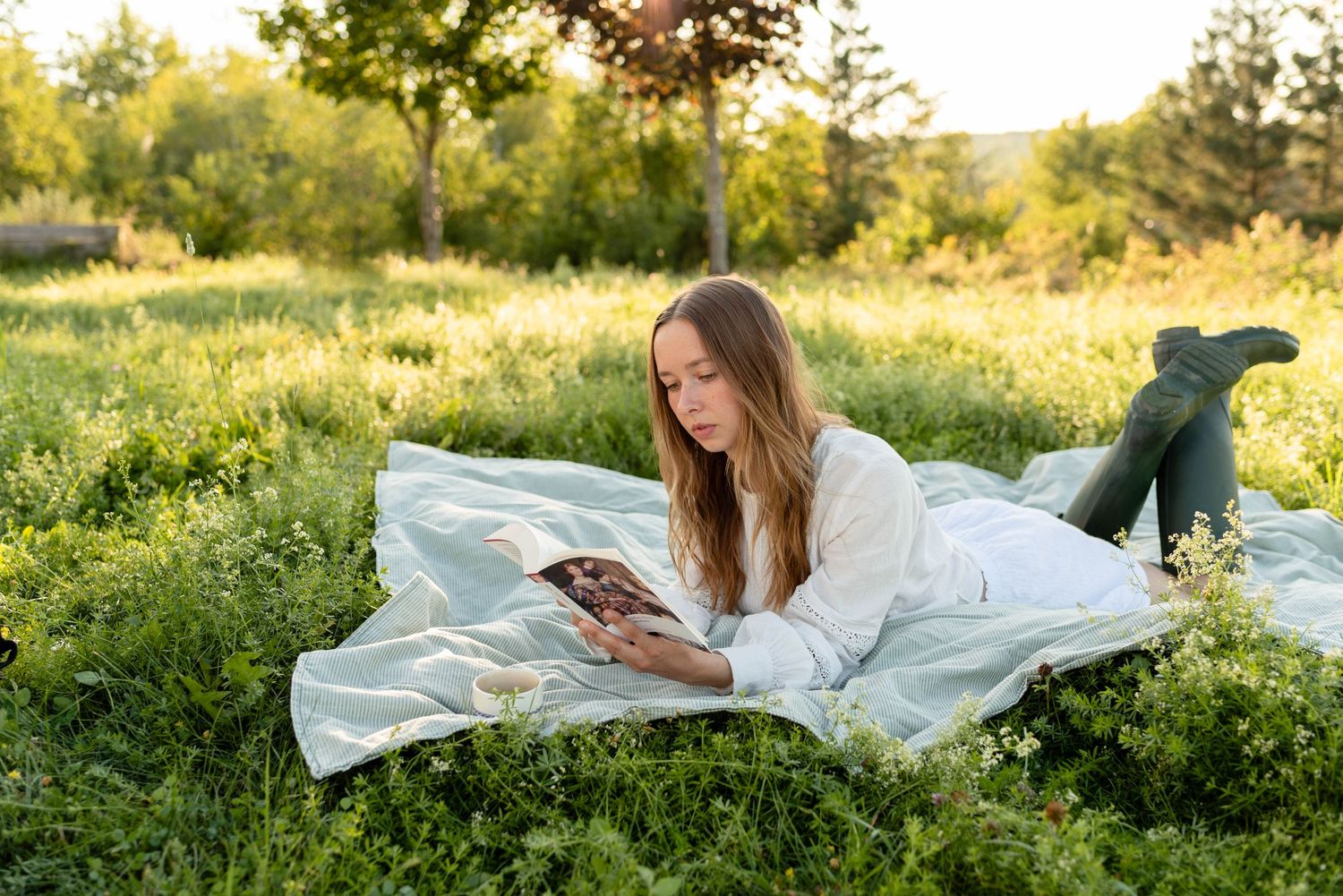Young woman relaxing on white blanket in lush green grass reading a book at sunset.