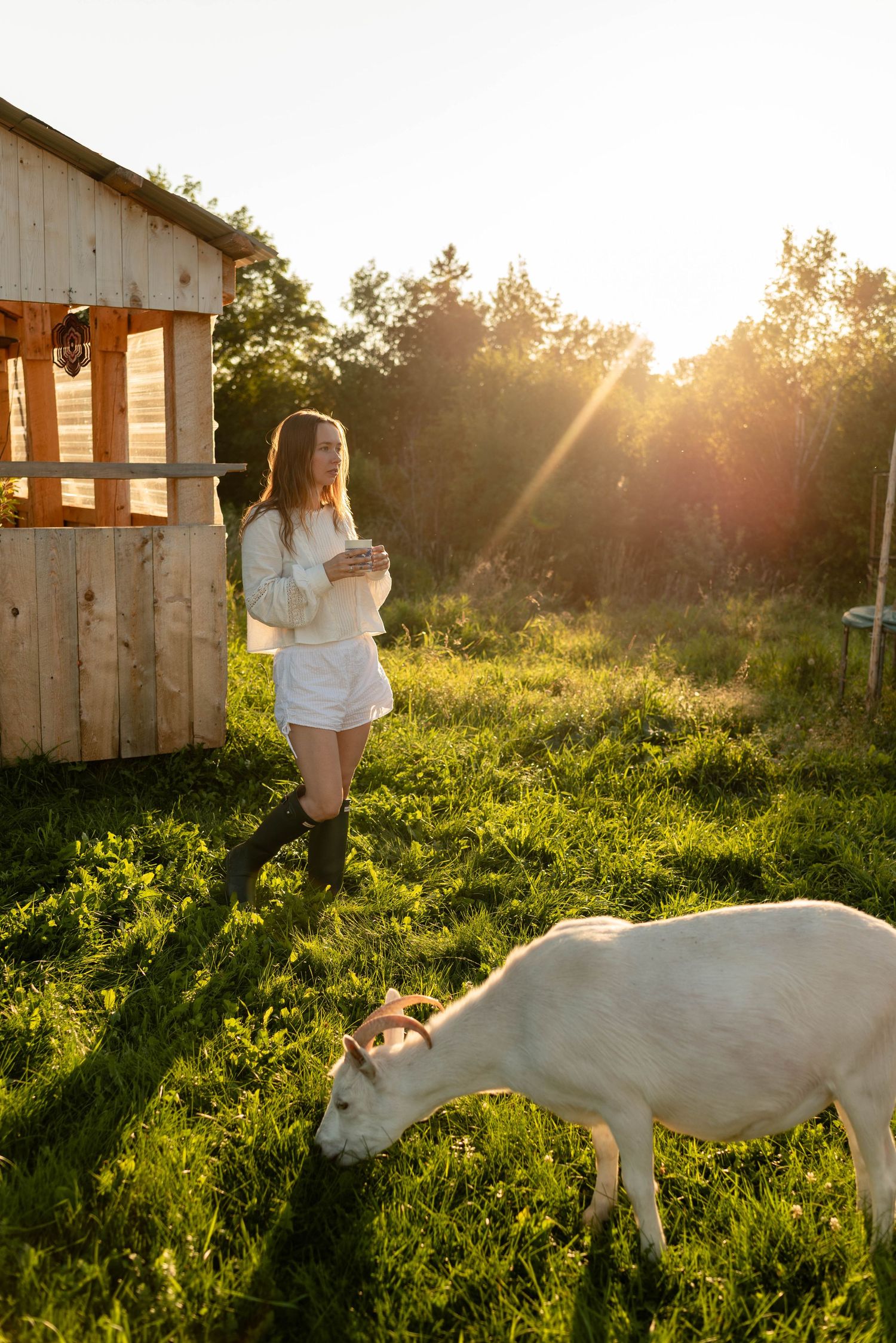 Woman in white outfit with rubber boots standing near wooden shed with white goat.