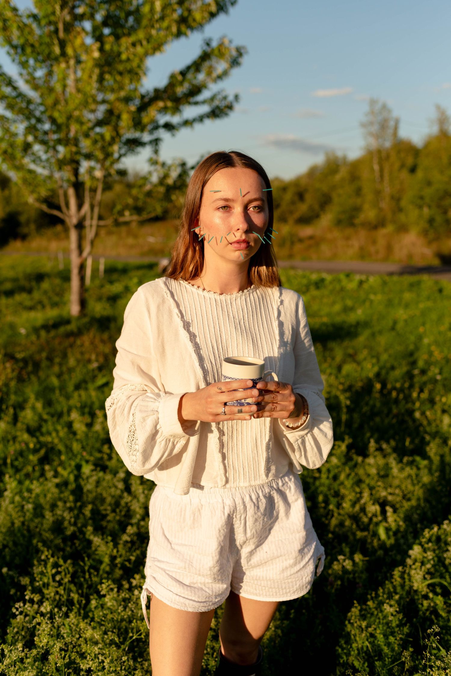 Young woman in white outfit holding coffee cup standing in sunny green field.