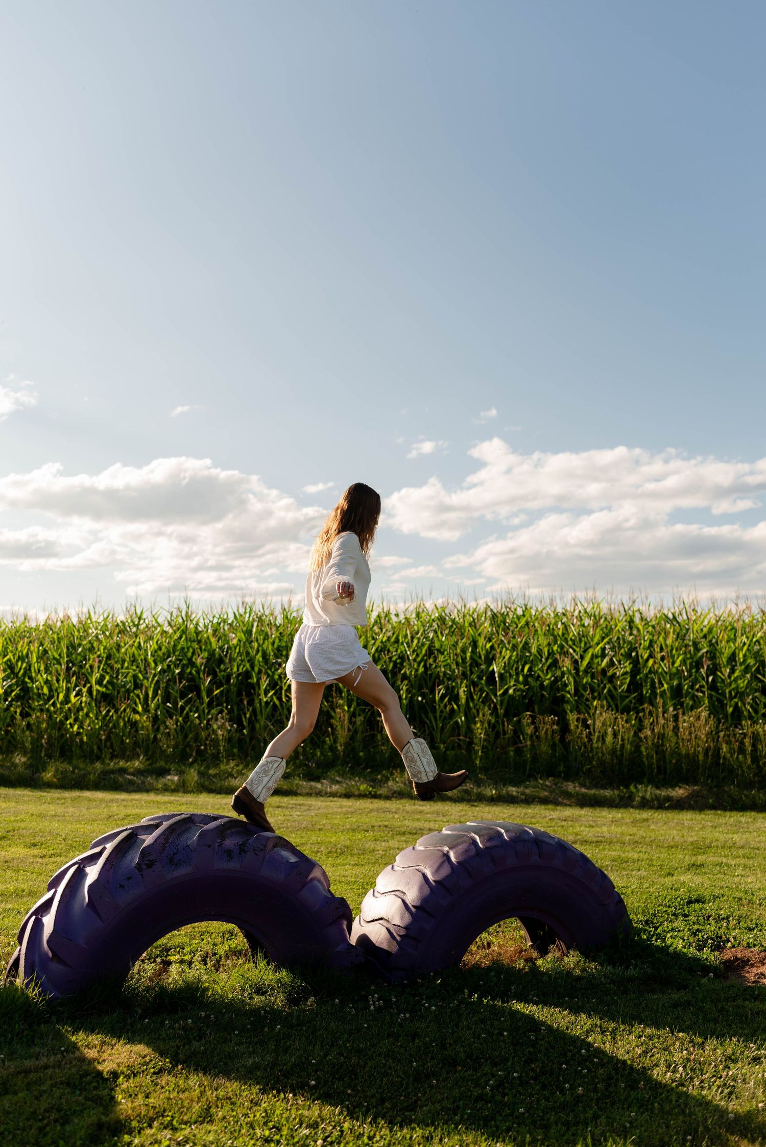 Woman in white shorts and cowboy boots stepping over large rubber tires near a cornfield outdoors.
