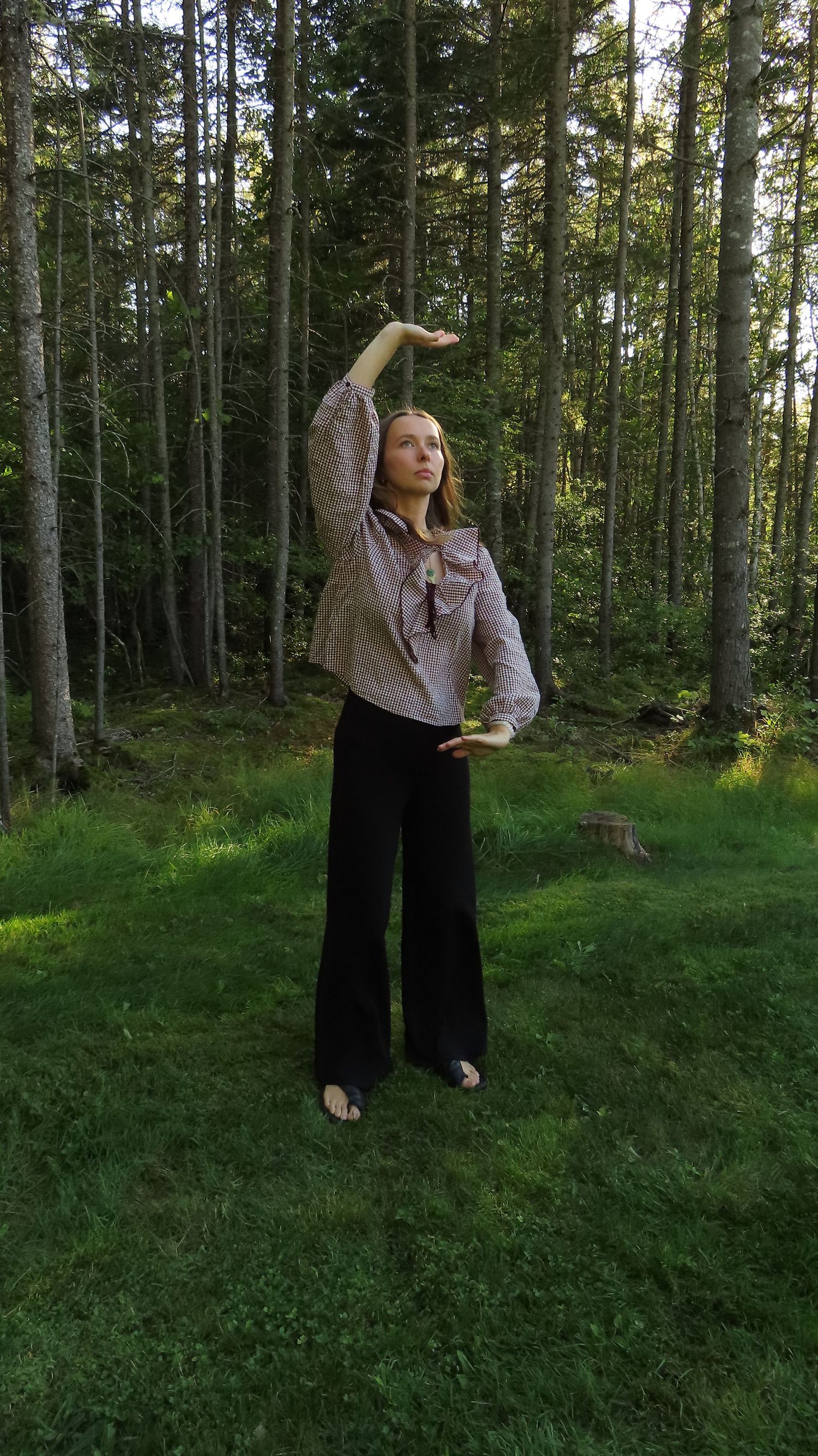 Young woman in ruffled gingham blouse posing gracefully with raised arm in a pine forest.