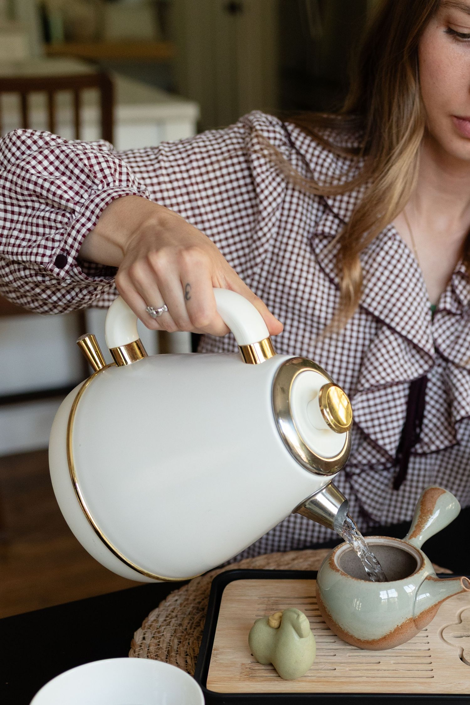 Woman in gingham blouse pouring hot water from white and gold kettle into ceramic teapot on wooden tray.