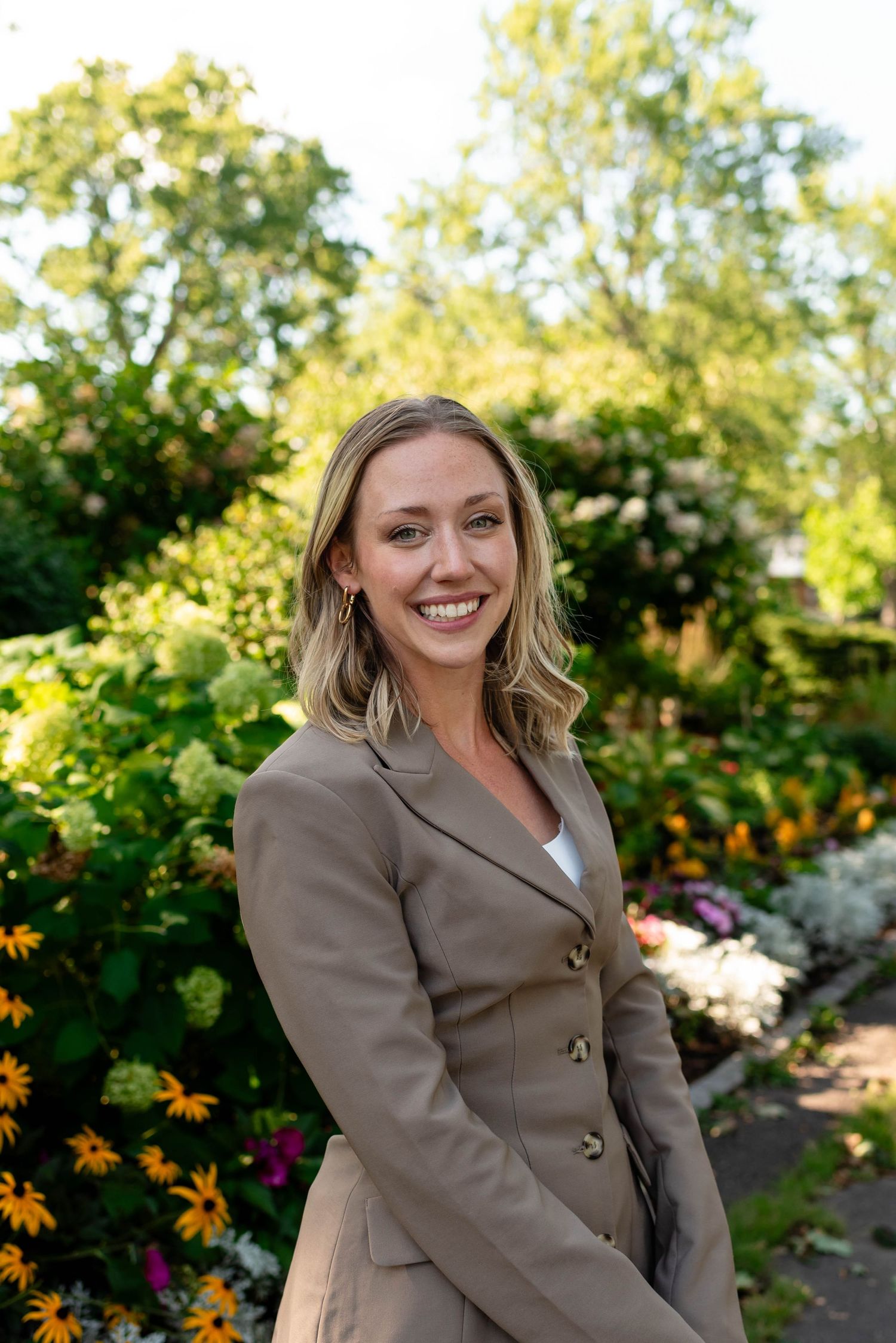 Smiling blonde woman in khaki blazer dress posing in a vibrant sunlit garden setting.