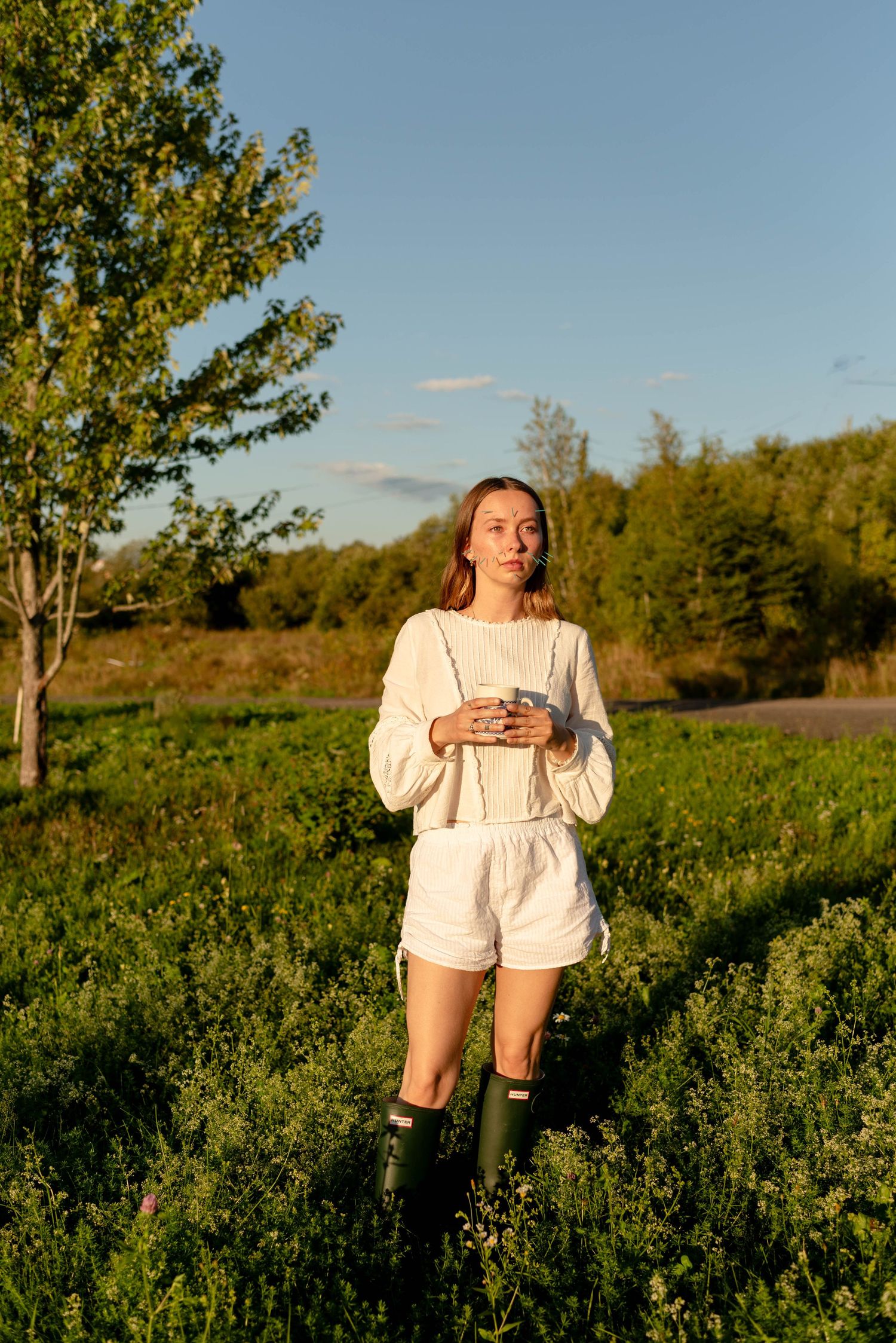 Young woman in white outfit and green boots standing in sunlit green field holding phone.