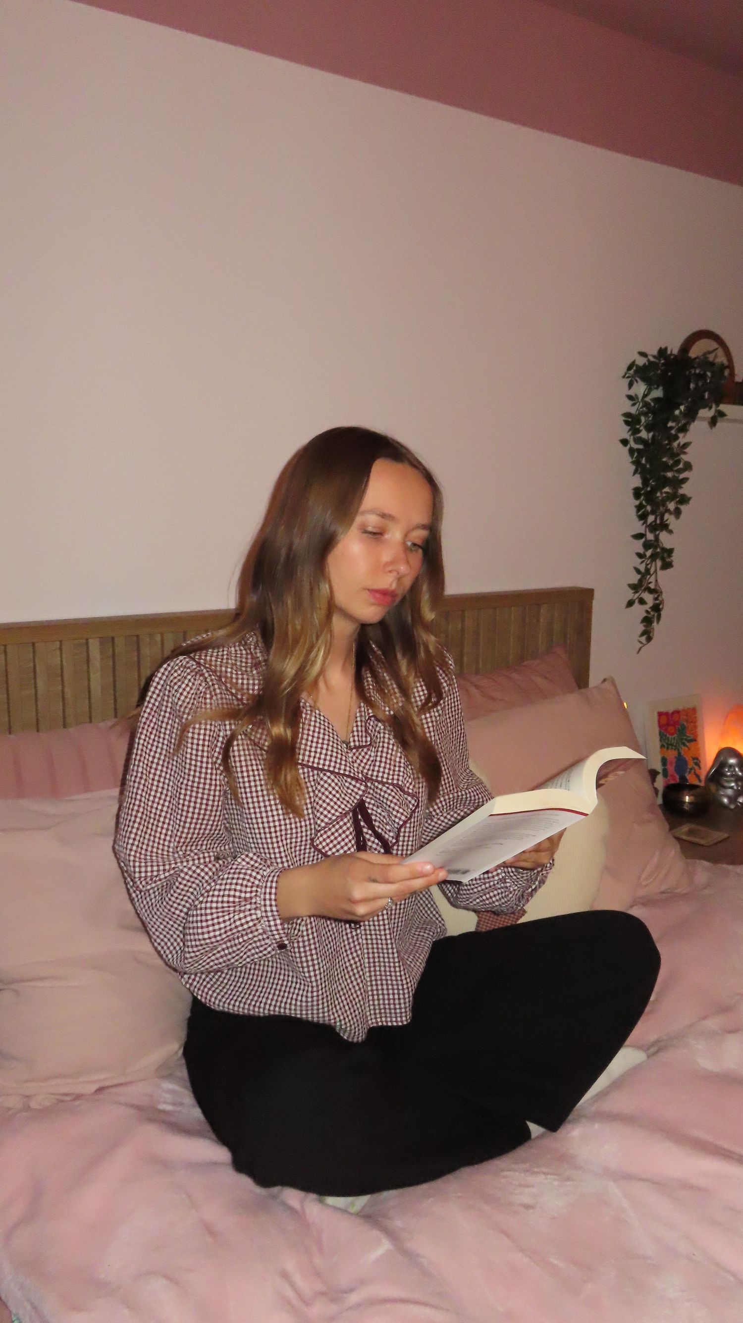 Young woman in gingham ruffle blouse sitting cross-legged on bed reading a book indoors.