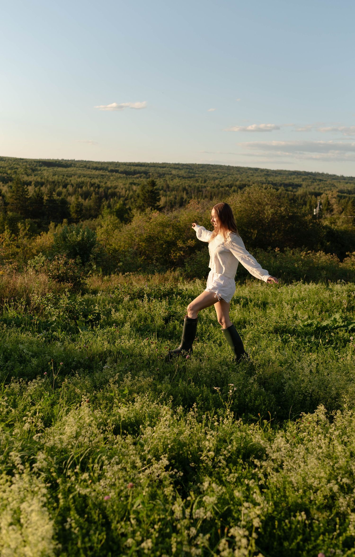 Young woman in white dress and black boots dancing in a lush green meadow at golden hour.