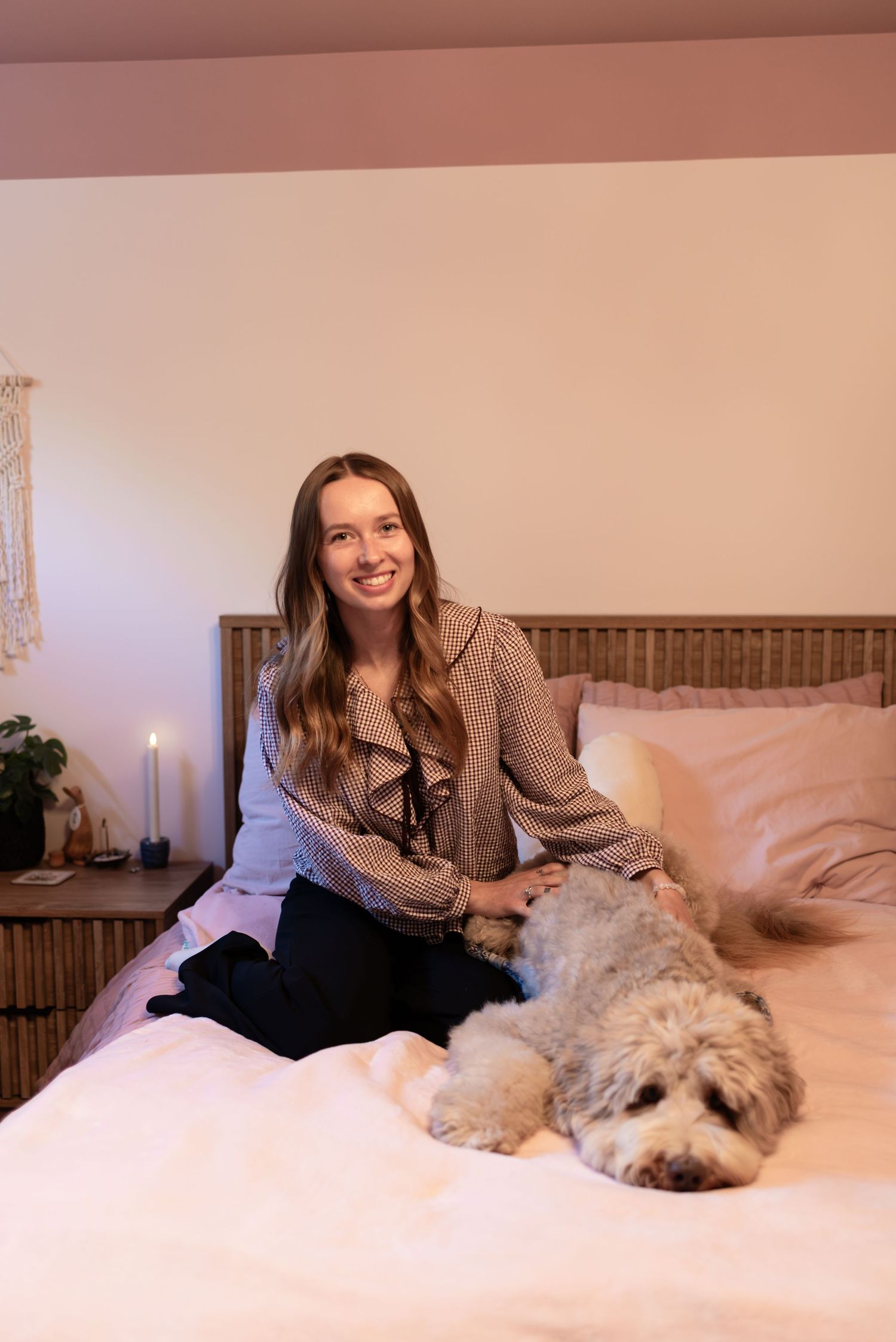 Smiling young woman sitting on bed with fluffy white dog in warmly lit bedroom with rattan headboard.