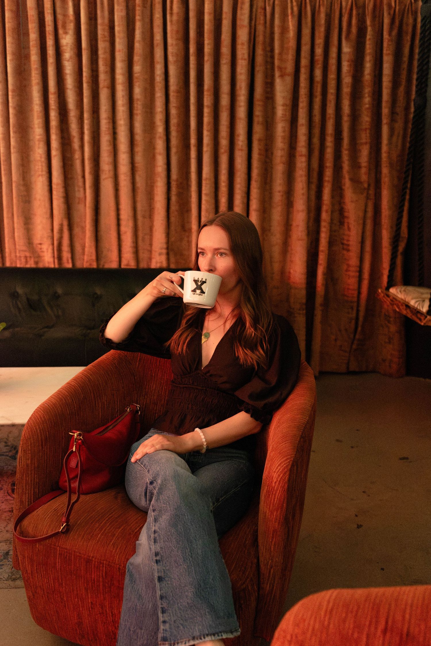 Woman in black top and jeans sipping from a mug in a red velvet armchair with warm ambient lighting.