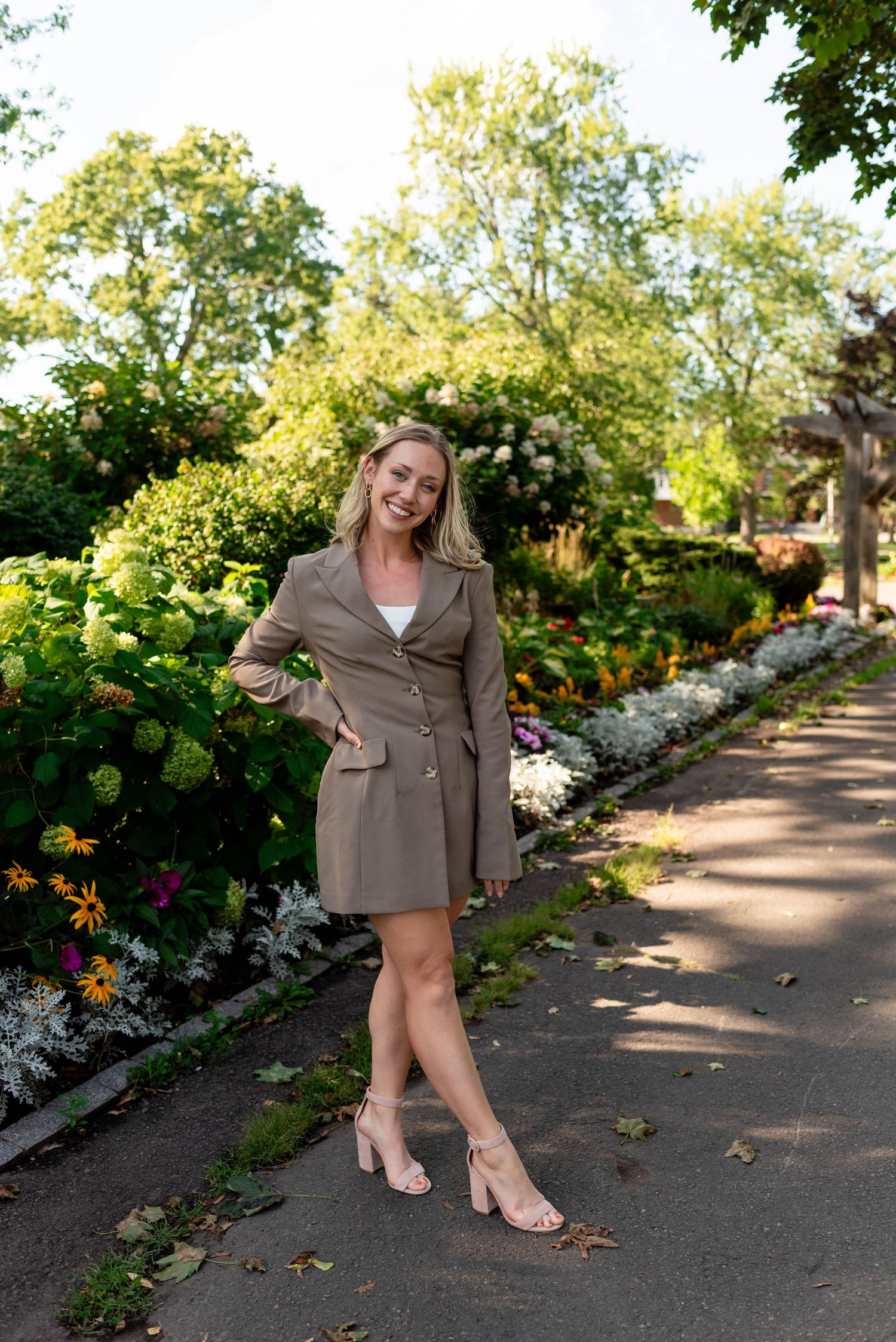 Confident blonde woman in olive blazer dress posing on a garden path surrounded by colorful flowers.