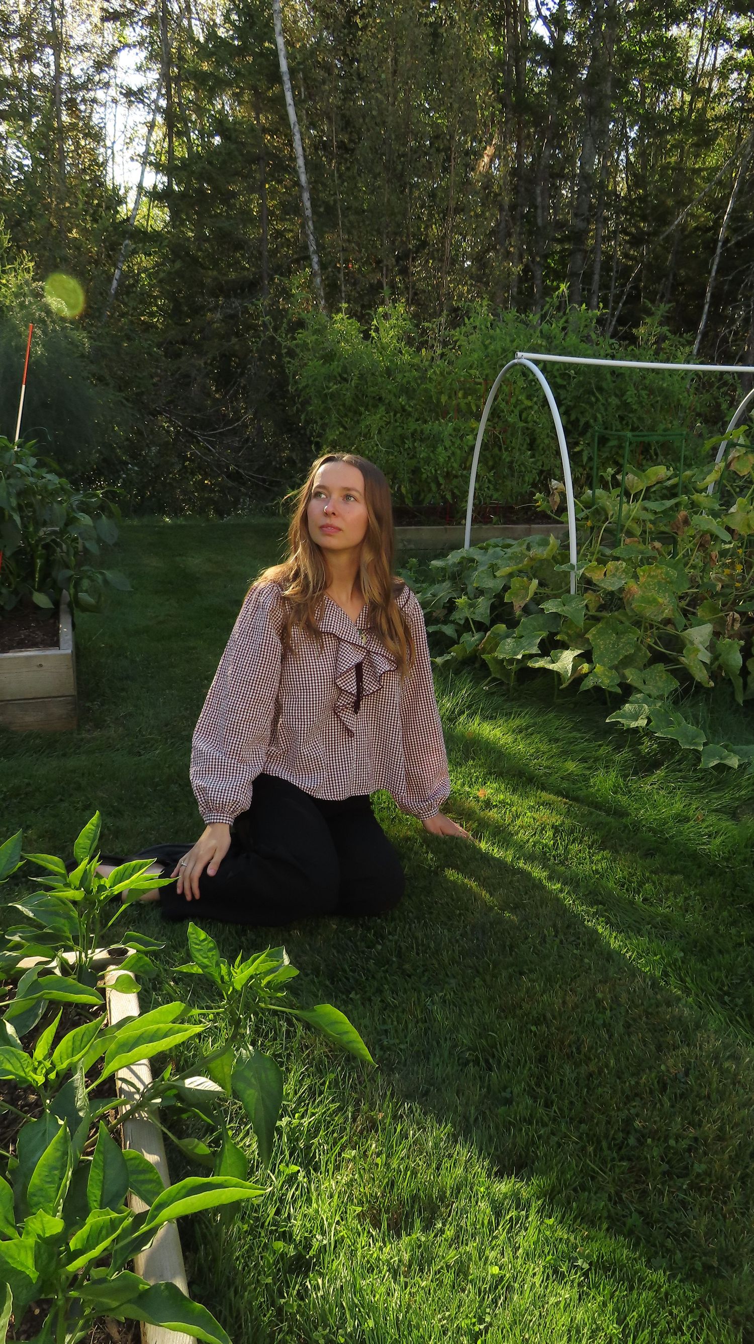 Young woman in pink ruffled blouse and black pants kneeling on lush green garden lawn surrounded by plants.