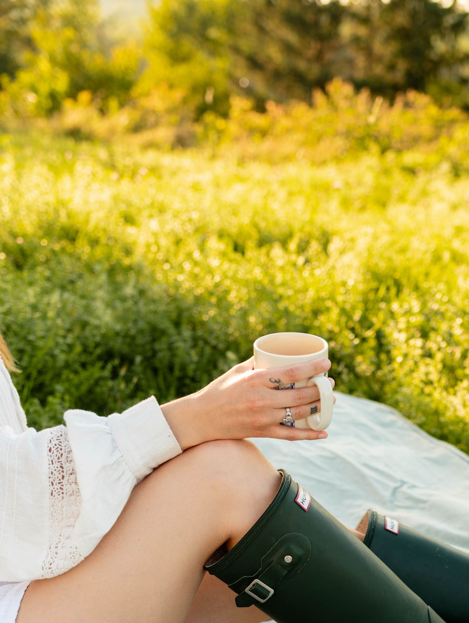 Woman in white lace top and green Hunter boots holding a ceramic mug outdoors on a blanket.
