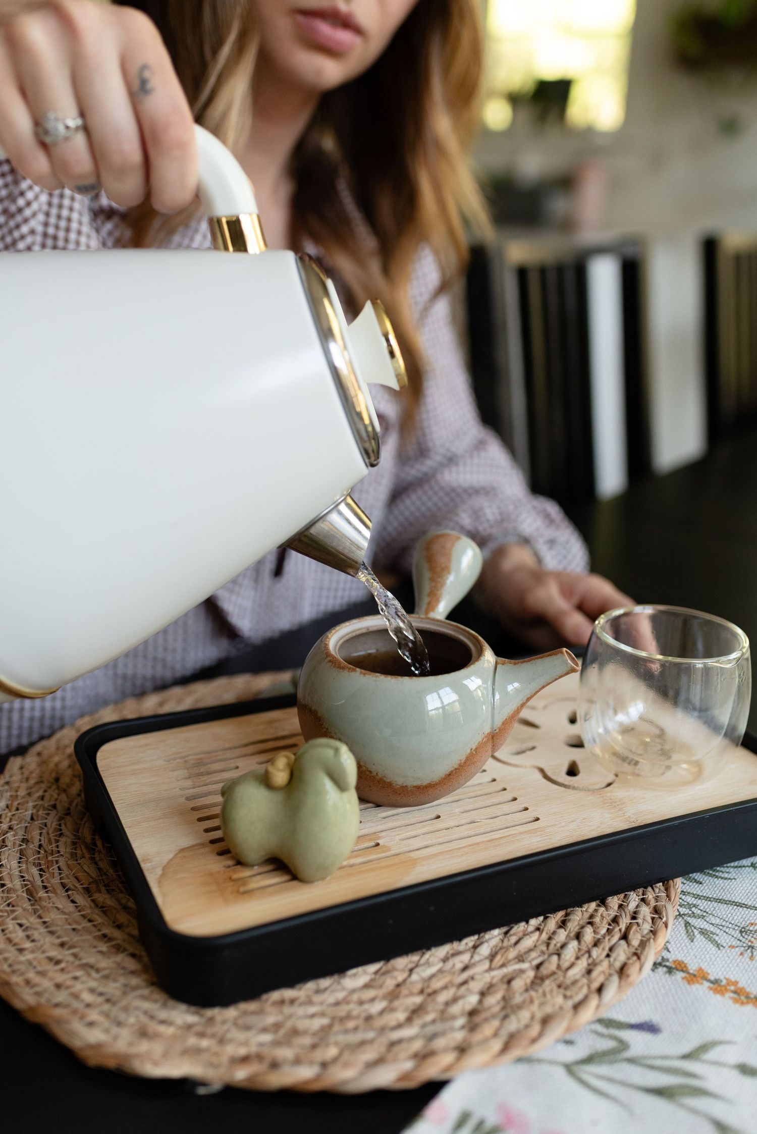 Woman pouring hot water from white kettle into ceramic teapot on wooden gongfu tea tray.