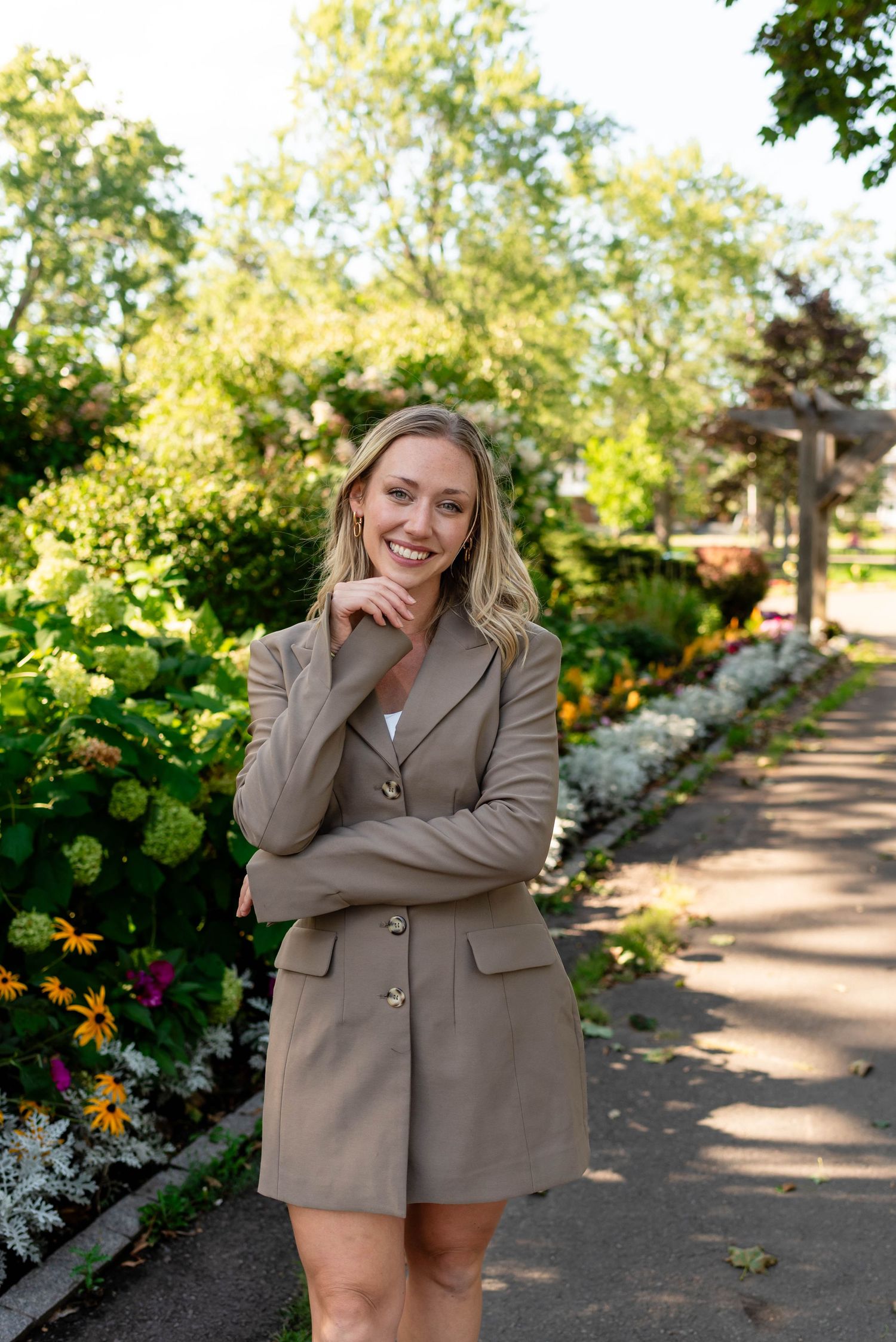 Happy blonde woman in tan blazer dress standing on garden path with arms crossed.