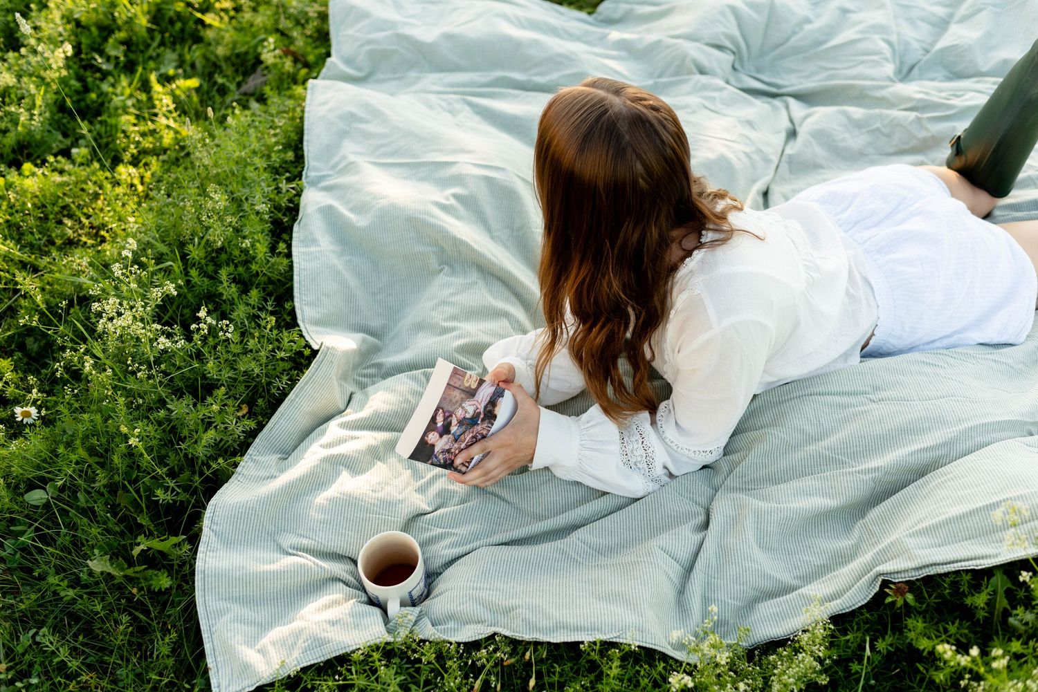 Woman lying on light blue blanket in grass reading a book with a cup of coffee nearby.