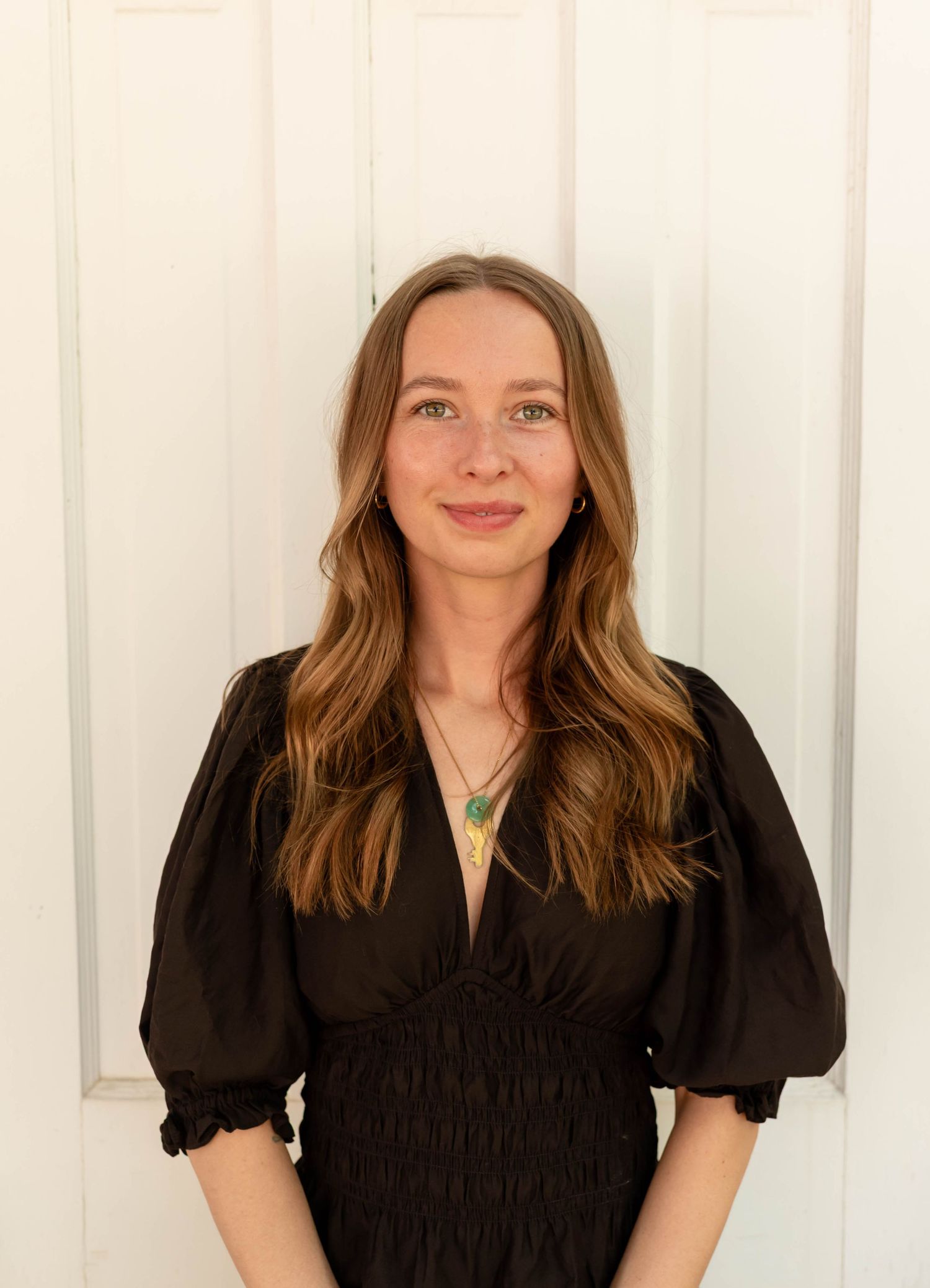 Young woman with long brown hair in black dress wearing green pendant necklace against white door.